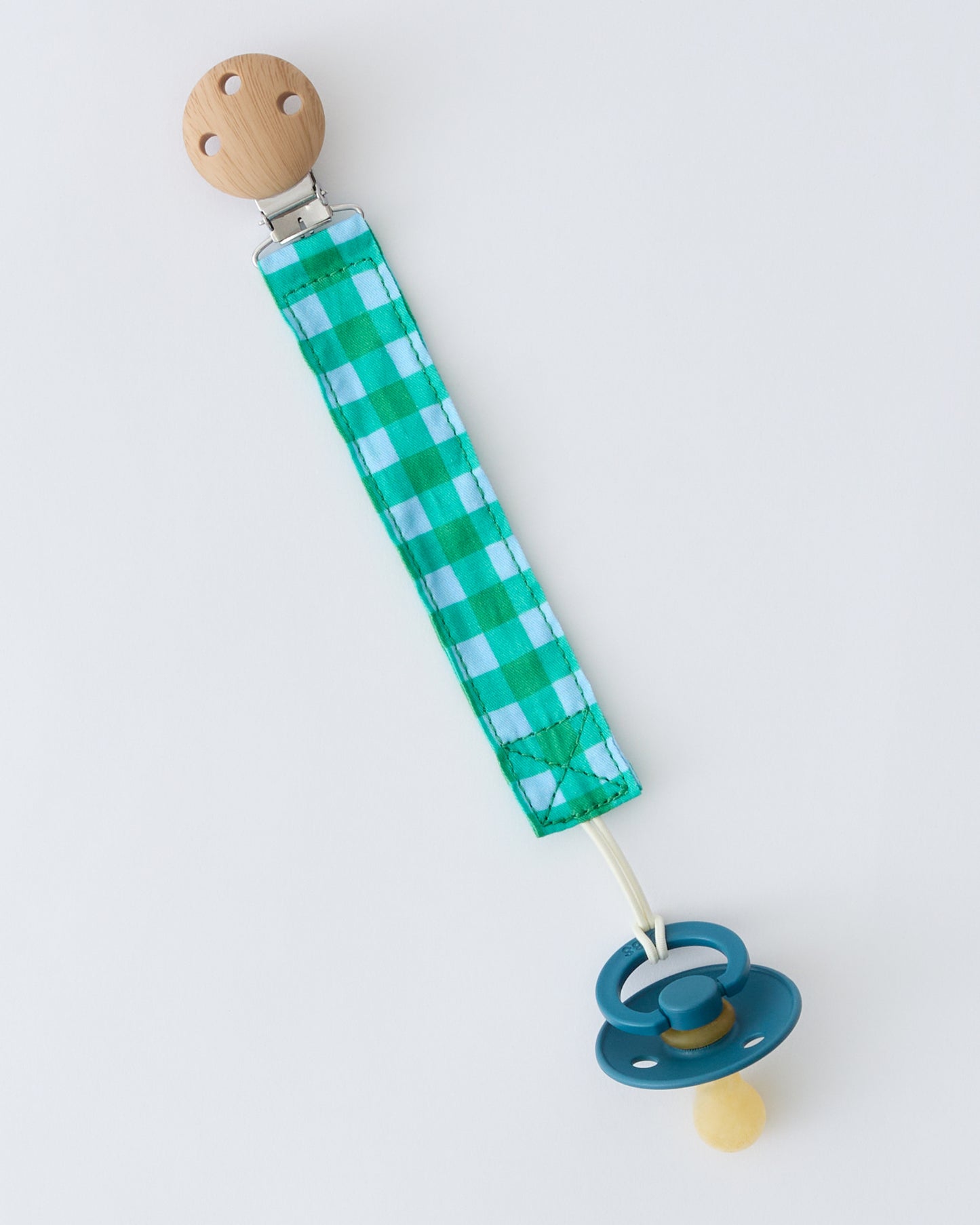 Pacifier Clip Two-Pack in Teal Gingham, attached to pacifier, on a beige background.