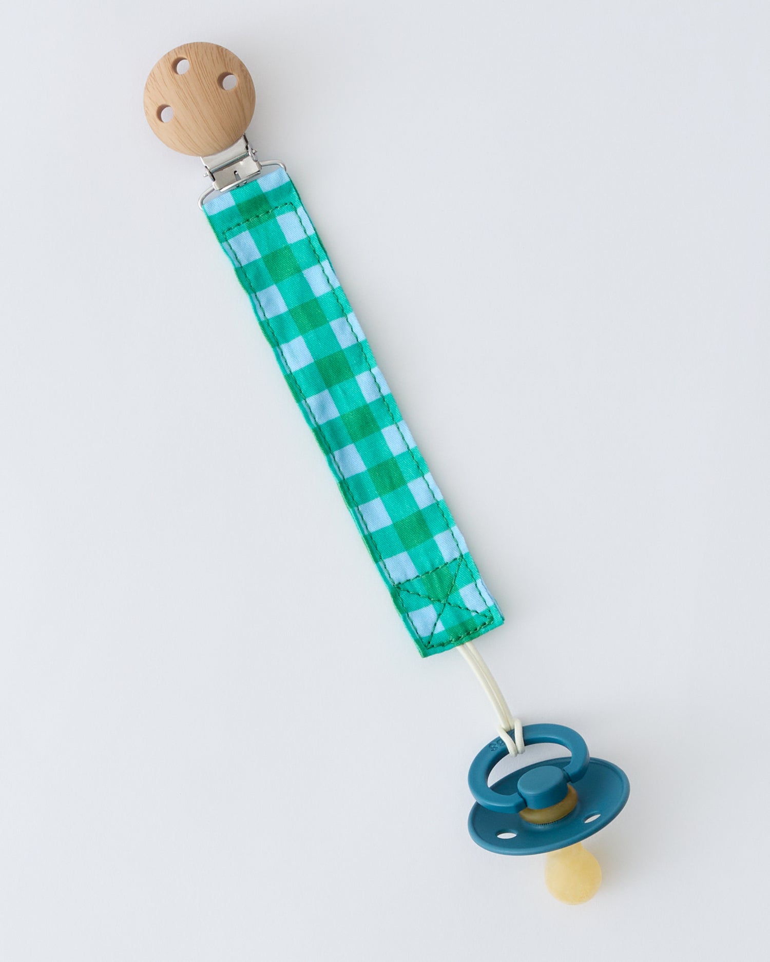 Pacifier Clip Two-Pack in Teal Gingham, attached to pacifier, on a beige background.