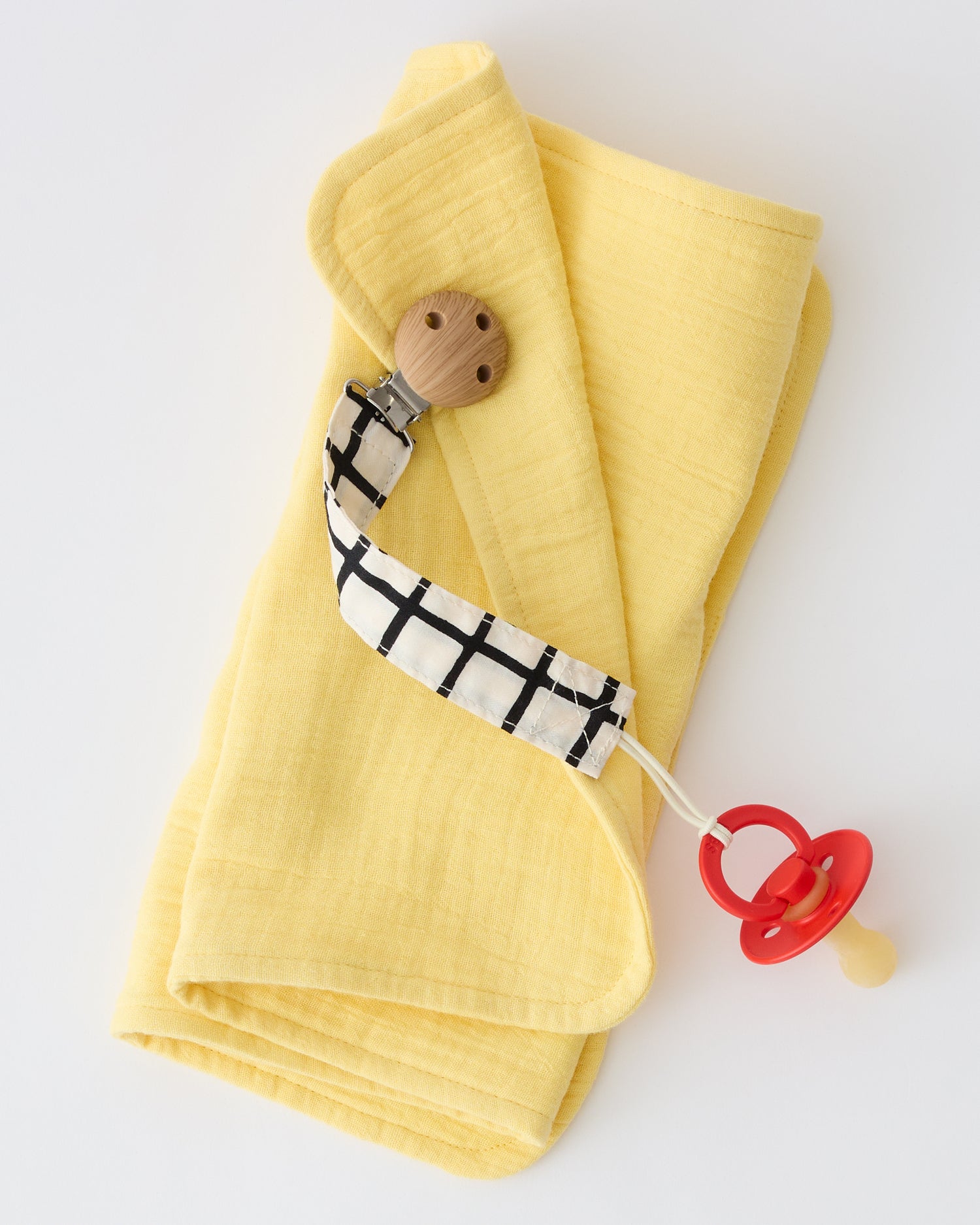 Pacifier Clip Two-Pack in Black Grid, holding onto a pacifier and clipped to a muslin cloth, on a beige background.