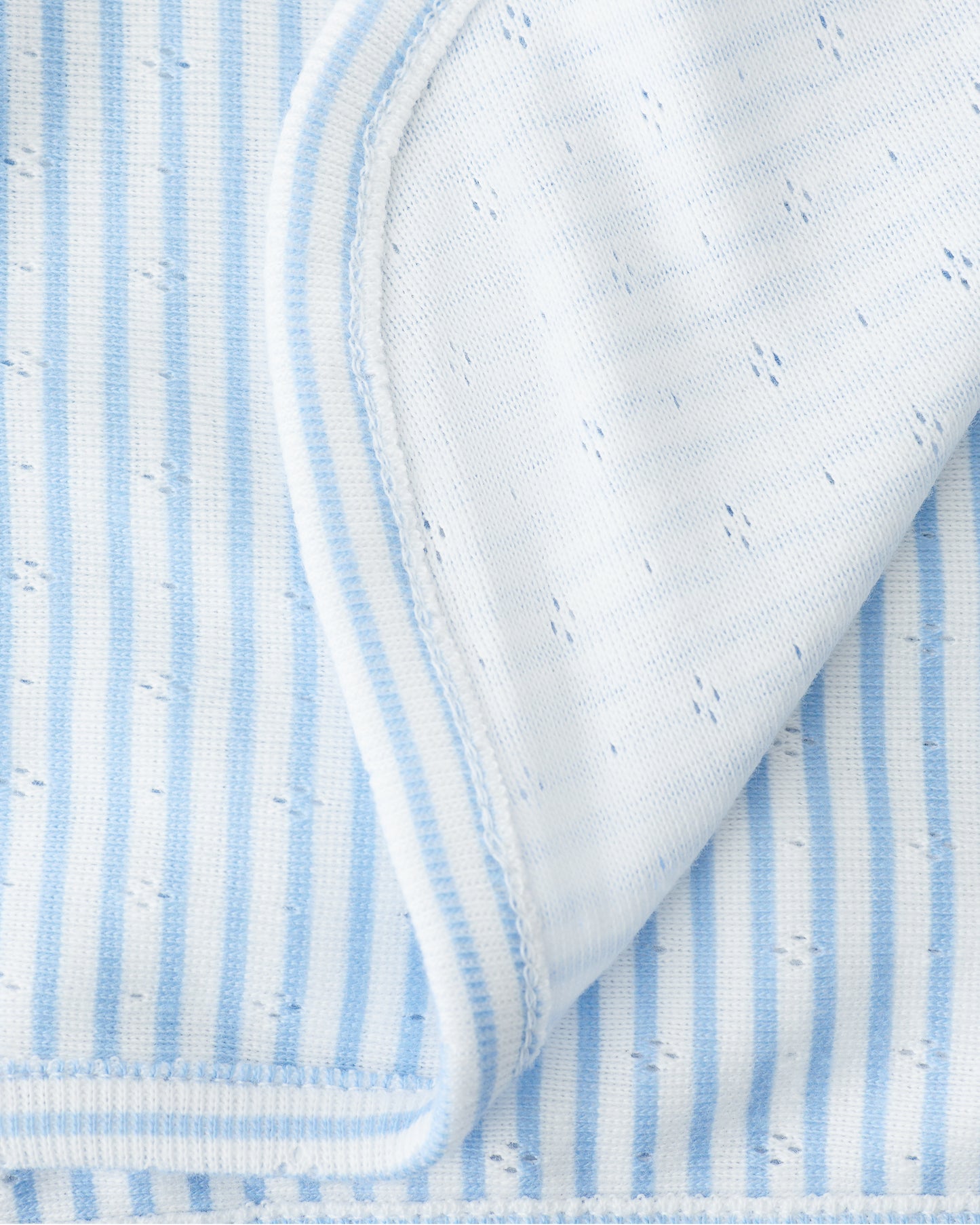 Close up of the Pointelle Swaddle in Blue Candy Stripe folded with a flipped corner to show reverse side, on a beige background.