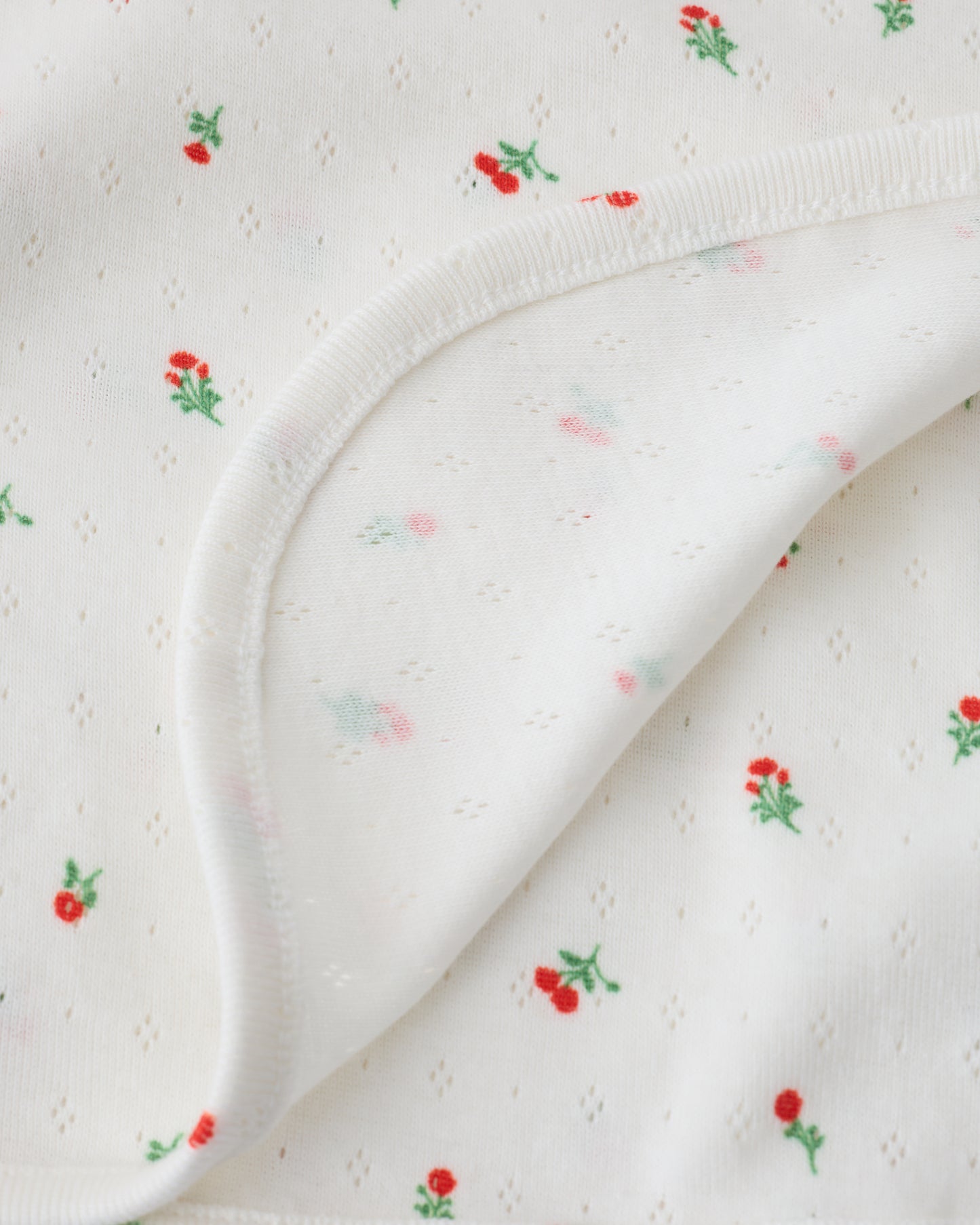 Close up of the Pointelle Swaddle in Dainty Ditsy pattern folded with a flipped corner to show reverse side, on a beige background.