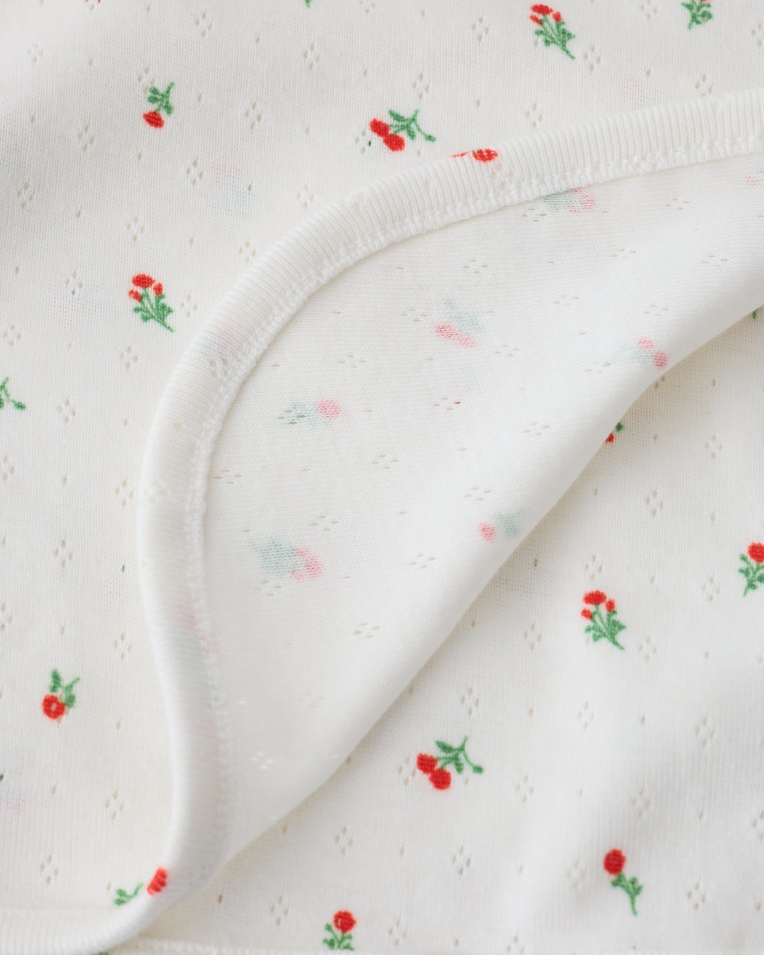 Close up of the Pointelle Swaddle in Dainty Ditsy pattern folded with a flipped corner to show reverse side, on a beige background.