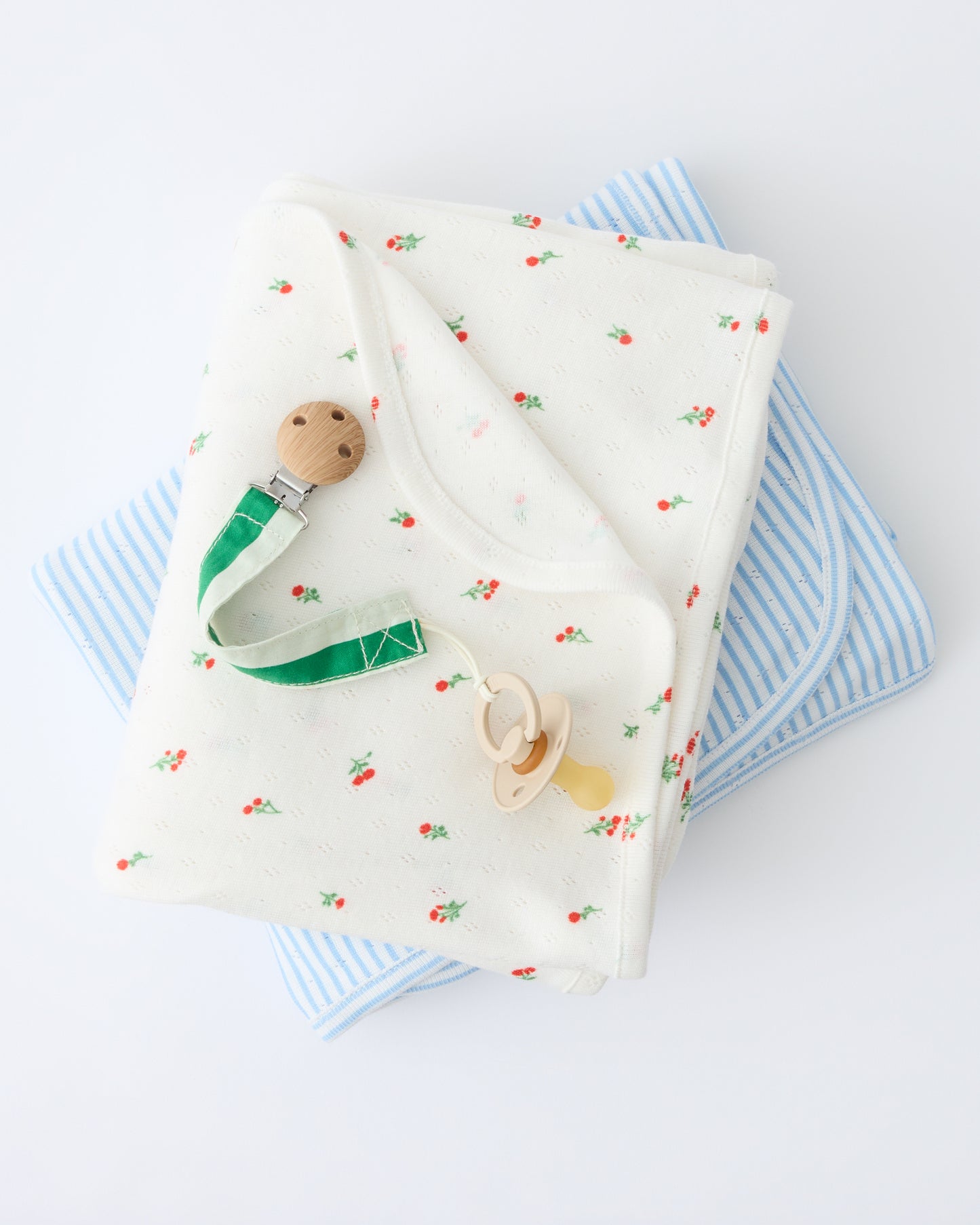 Pointelle Swaddle in Dainty Ditsy pattern and Blue Candy Stripe are folded and stacked with a flipped corner to show reverse side, with a Pacifier Clip in Green Stripe, on a beige background.