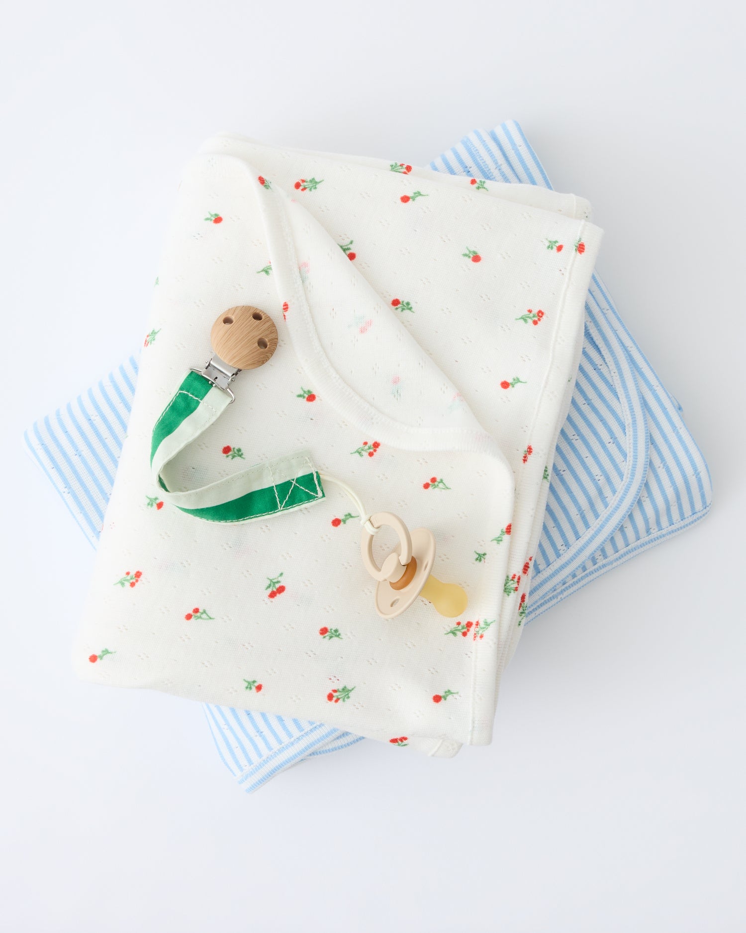 Pointelle Swaddle in Dainty Ditsy pattern and Blue Candy Stripe are folded and stacked with a flipped corner to show reverse side, with a Pacifier Clip in Green Stripe, on a beige background.