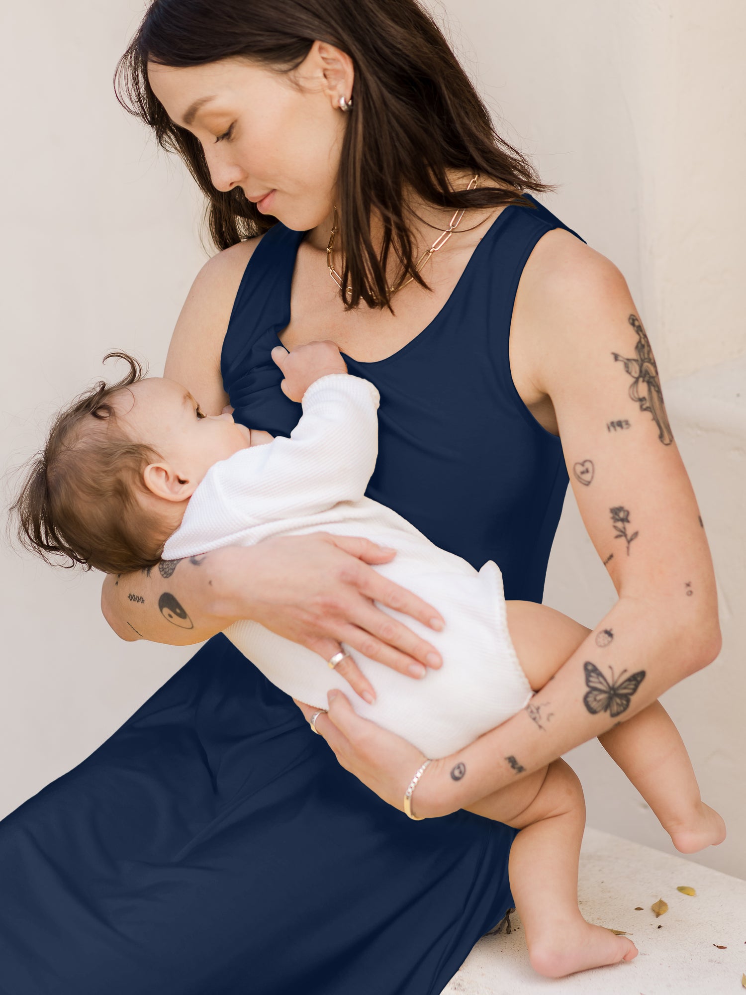 Model sitting down wearing the Penelope Crossover Nursing Dress in navy and nursing baby
