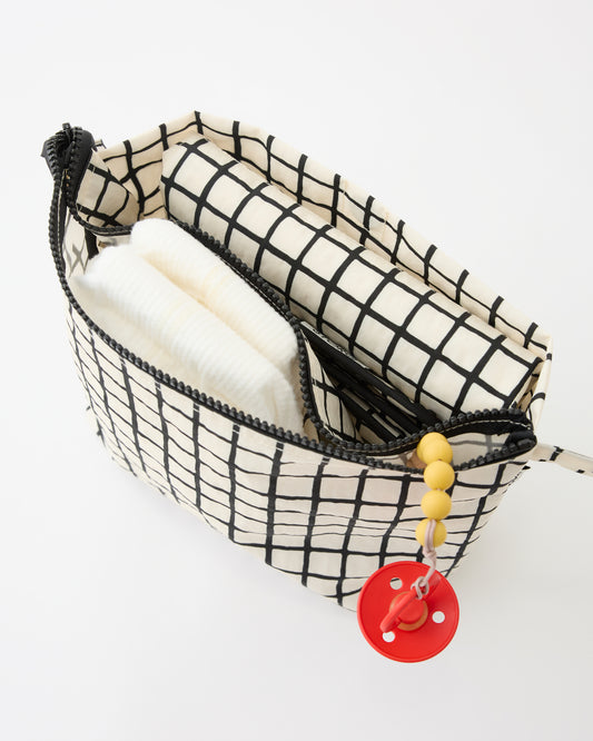 Close up of diaper, pacifier and folded mat inside pocket of the Quick Change Kit Bag in Black Grid, on a beige background.