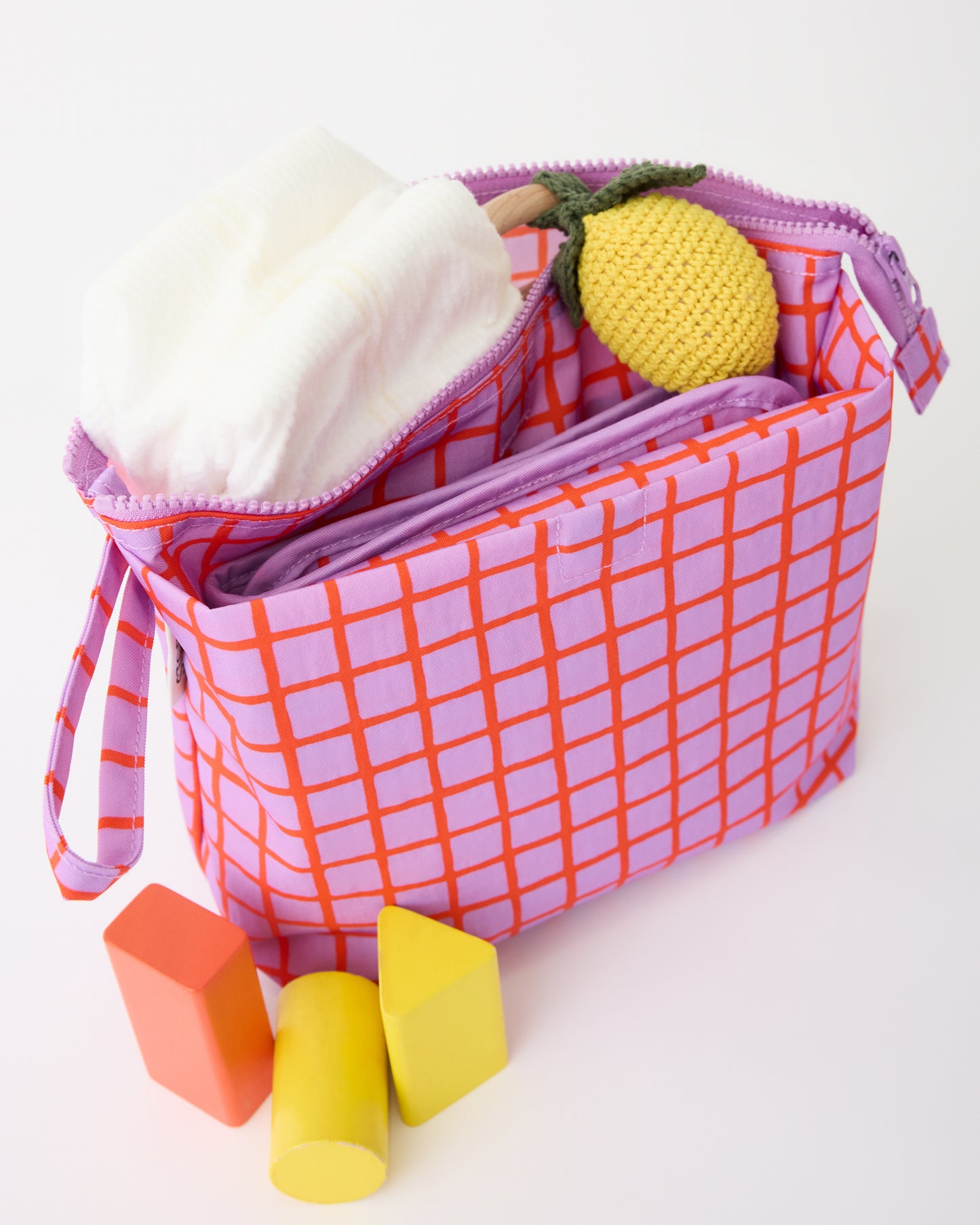 Quick Change Kit Bag in Orchid Grid, with diapers, lemon lovey and changing mat sticking out of pocket, and toys on floor beside it, on a beige background.