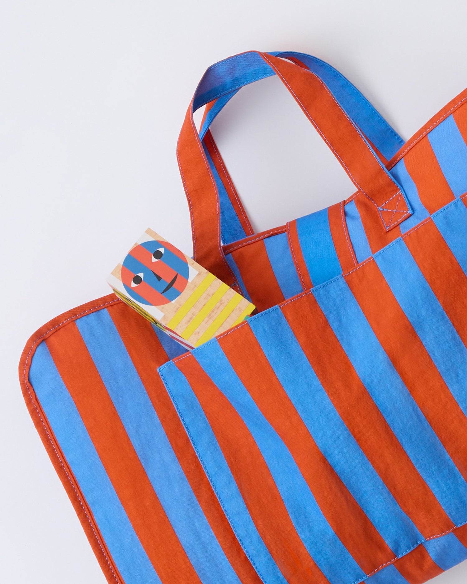 The Cool Kid Nap Mat in Blue & Rust Stripe folded up and ready to go, with a little toy sticking out of the outer pocket, on a beige background.