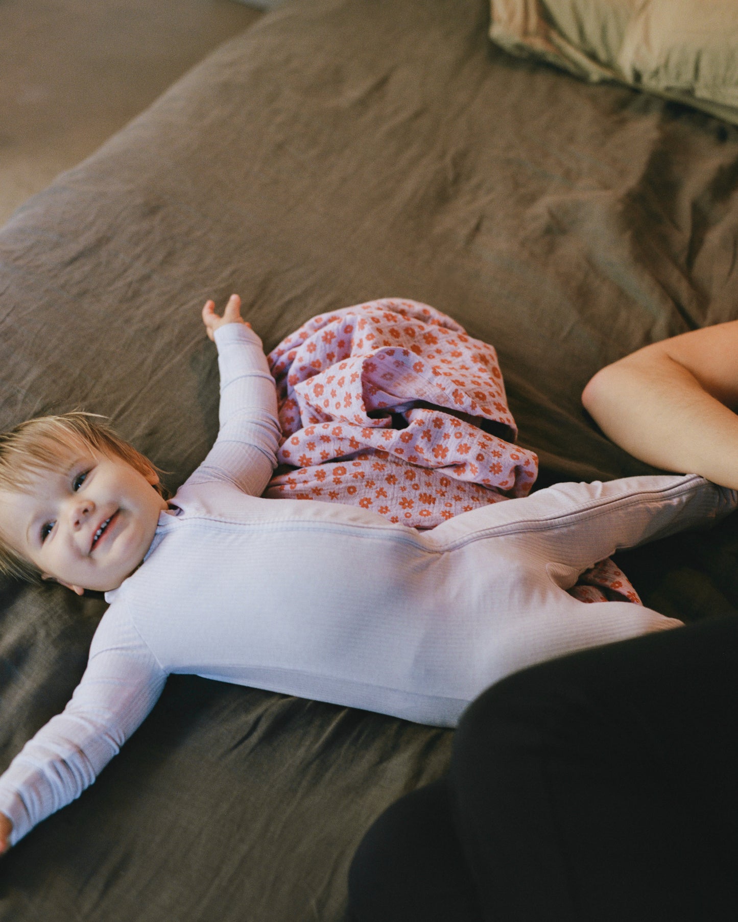 A baby lying on a bed, smiling, next to the Organic Muslin Swaddle in the Lavender Cosmos print.@model_info:Palma is wearing 12-18 Months.