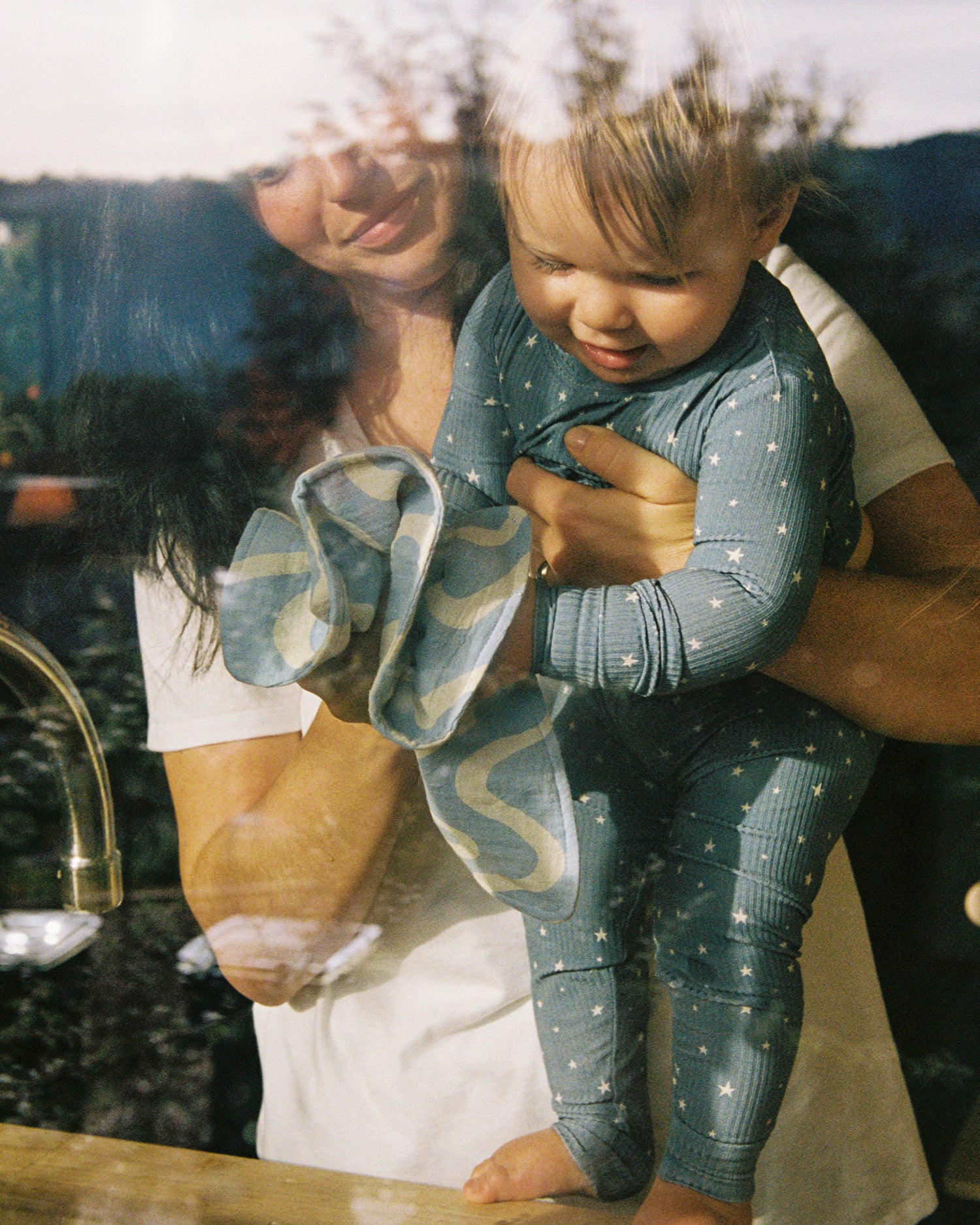 A woman holding a baby playing with the Organic Muslin Burp Cloth in the Blue Waves print.