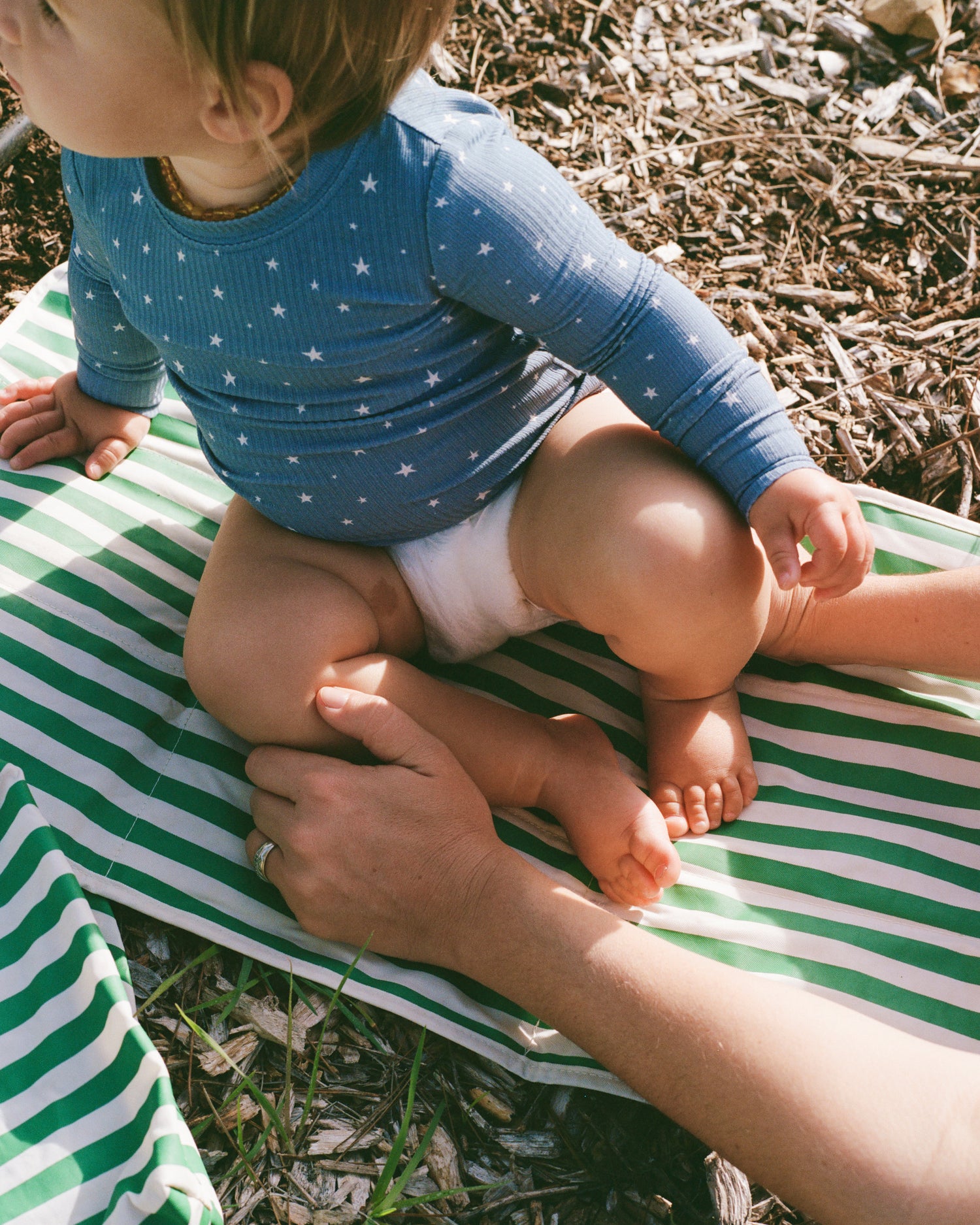 Baby wearing blue star pattern ribbed two piece pajama top sitting on a green and white striped blanket outdoors.