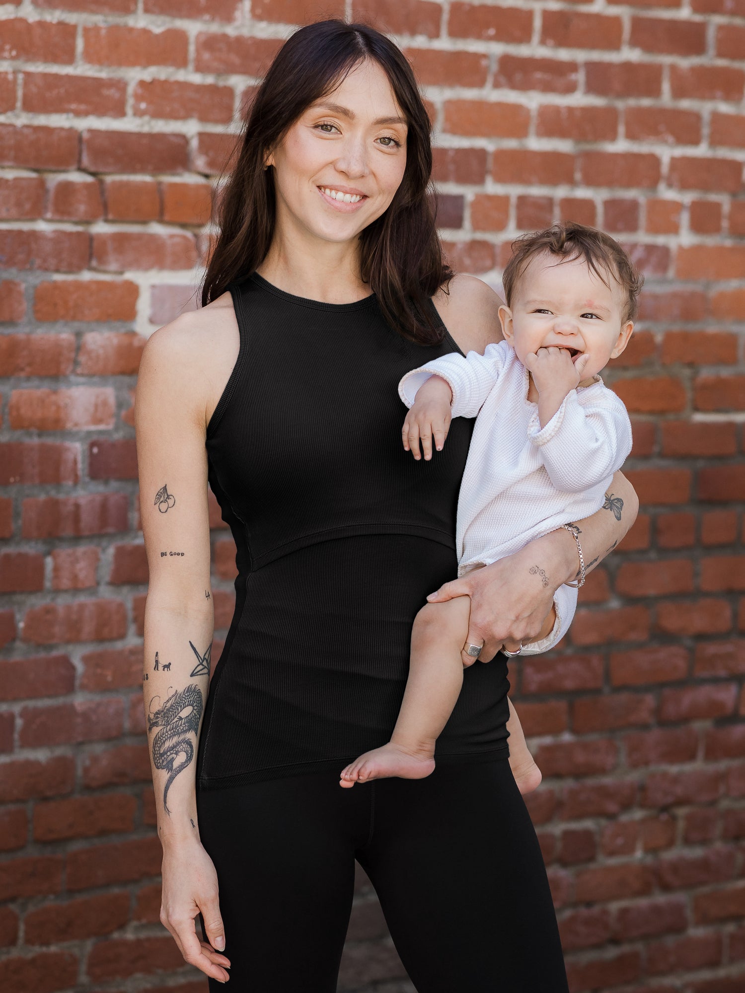 Front view of model holding baby and wearing the Ribbed Bamboo Racerback Nursing Tank in black @model_info:Lyn is wearing a Small.