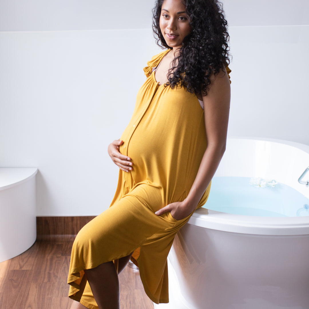 Pregnant model sitting on edge of bath tub wearing the Ruffle Strap Labor & Delivery Gown in honey
