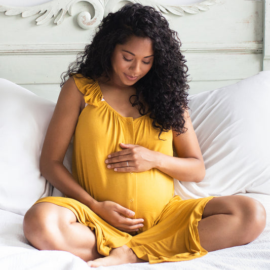 Pregnant model sitting on bed wearing the Ruffle Strap Labor & Delivery Gown in honey