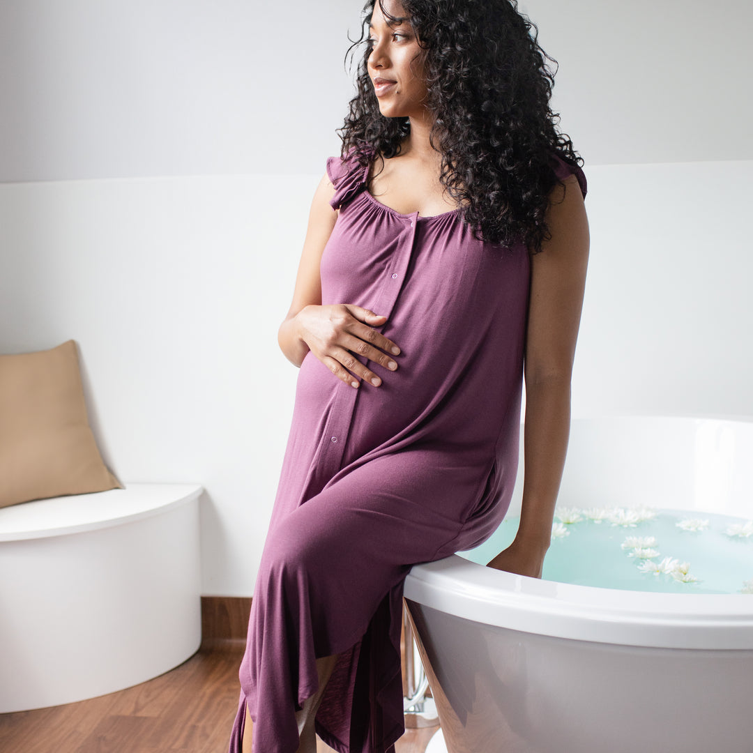 Model sitting on edge of bath tub wearing the Ruffle Strap Labor & Delivery Gown in Burgundy plum