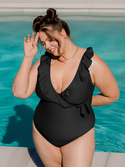 Model wearing the Ruffle Wrap Maternity & Nursing One Piece Swimsuit in Black with her hand on her hair and behind her back. @model_info:Bailey is wearing a Large Busty.