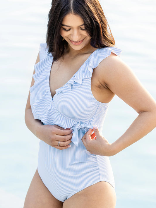 Model wearing the Ruffle Wrap Maternity & Nursing One Piece Swimsuit in Coastal Stripe with her hands on the tie. @model_info:Nohely is wearing a Medium.
