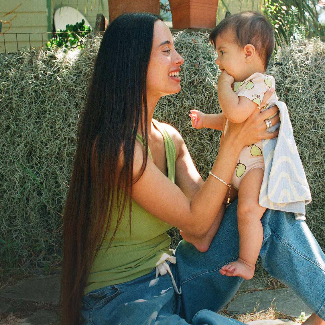 Woman holding baby outside, wearing a Pink Pomelo Onesie, with a Blue Wave Organic Muslin Burp Cloth, against a lush background.