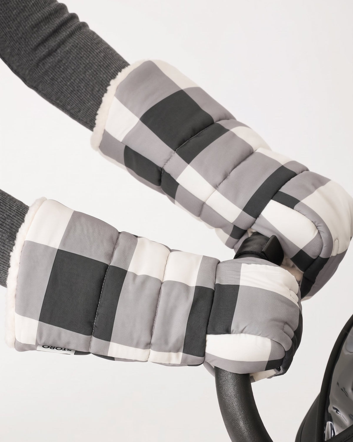 Model is wearing the Cozy Stroller Mitts in Buffalo Plaid while pushing the handlebar of a black stroller on an off white background.
