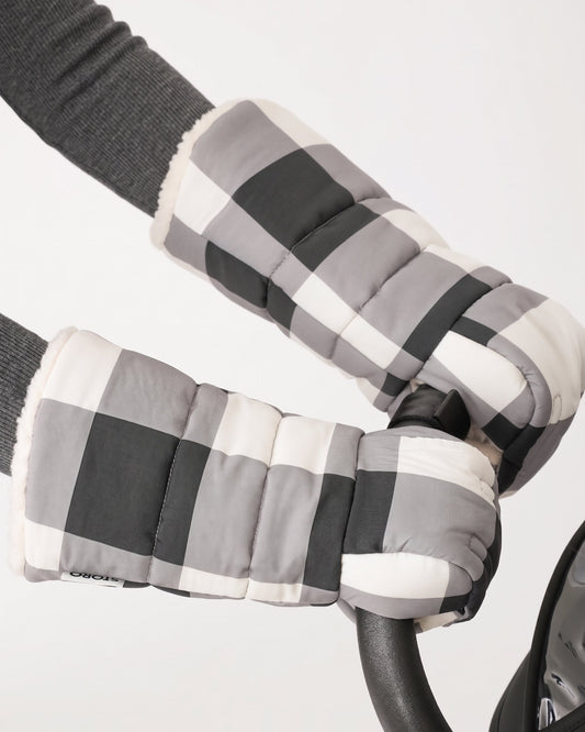 Model is wearing the Cozy Stroller Mitts in Buffalo Plaid while pushing the handlebar of a black stroller on an off white background.