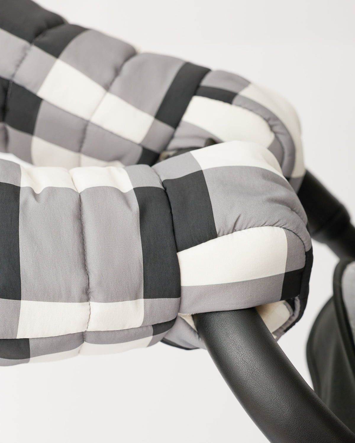 Model is wearing the Cozy Stroller Mitts in Buffalo Plaid while pushing the handlebar of a black stroller on an off white background.