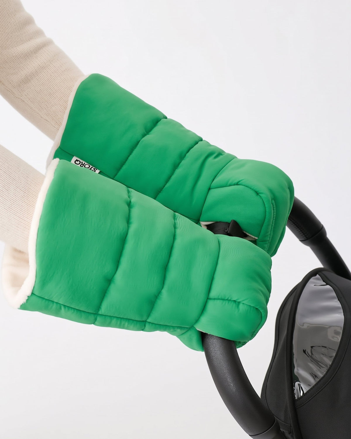 Model is wearing Cozy Stroller Mitts in Green while pushing a black stroller handlebar on an off white background.