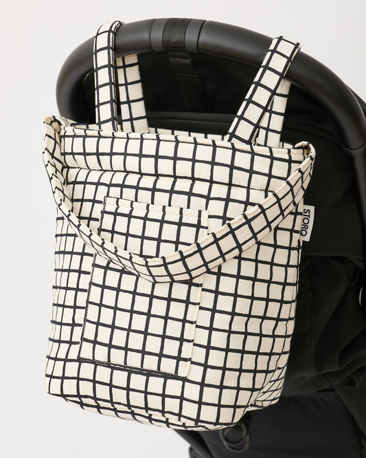 Black stroller with Stroller Party™ Bucket Bag in Black Grid attached to handlebar by the short handles, on an off white background.