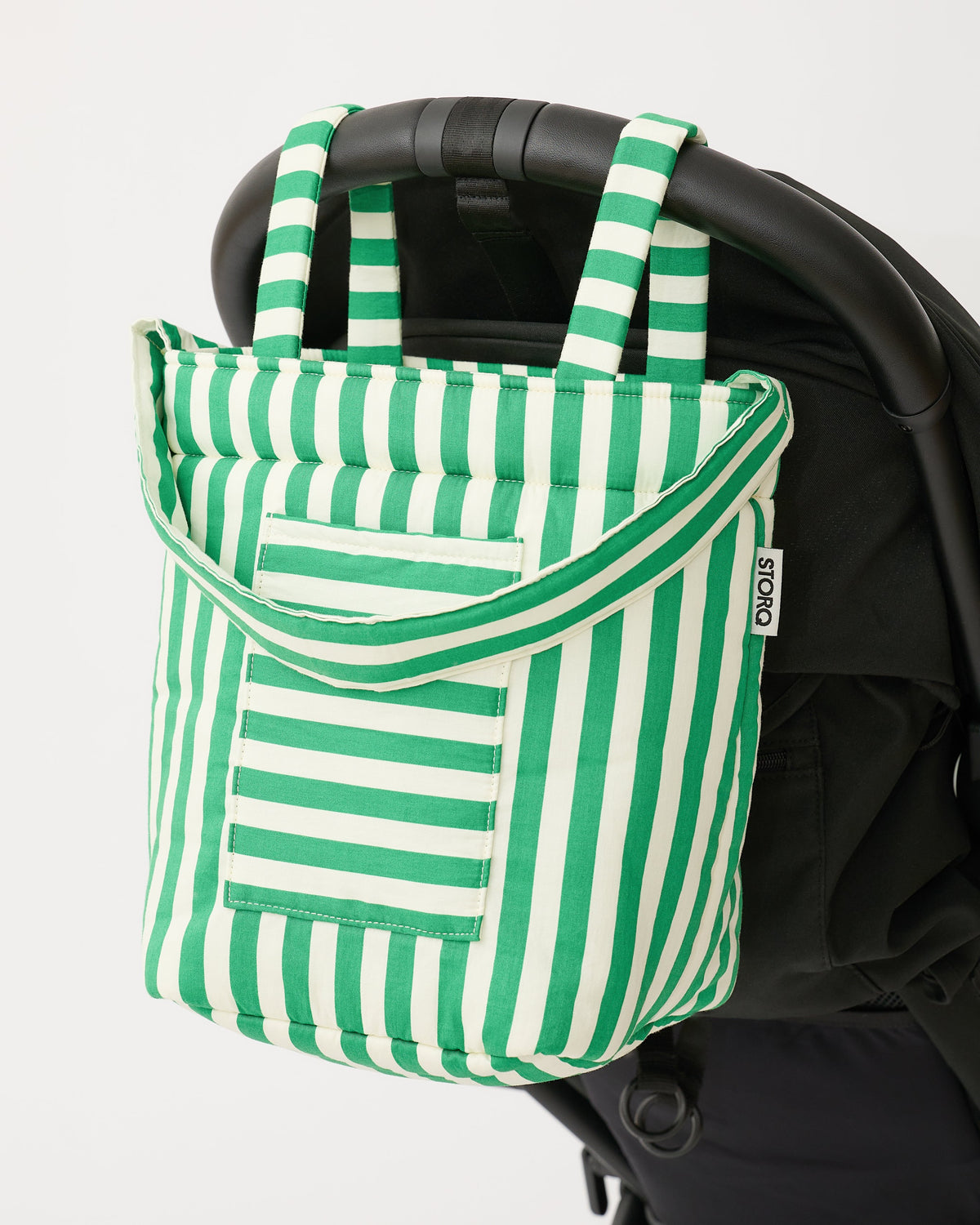 Stroller Party™ Bucket Bag in Green Stripe is hanging on handlebar of black stroller, on a beige background.