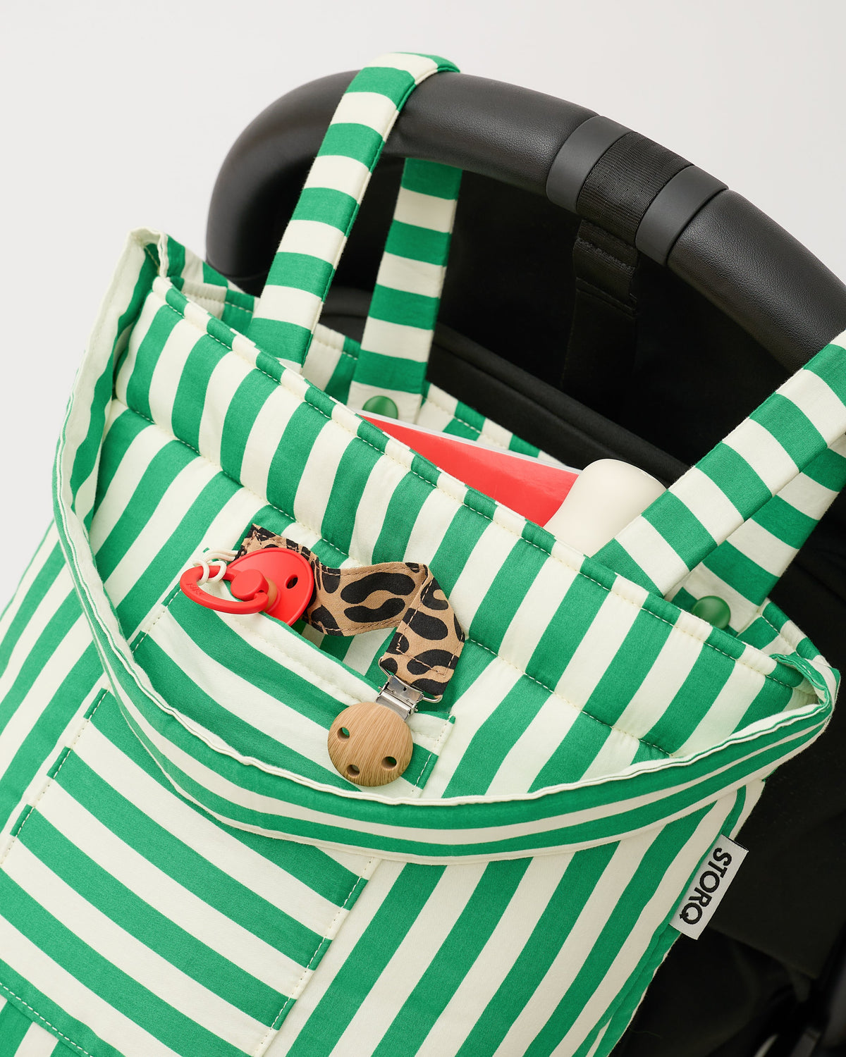 Stroller Party™ Bucket Bag in Green Stripe is hanging on handlebar of black stroller, with pacifier clip, bottle and other items peeking out, on a beige background.