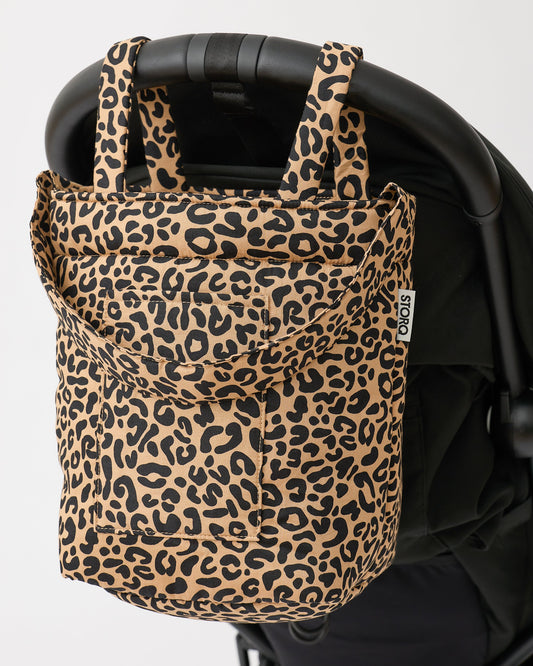 Stroller Party™ Bucket Bag in Leopard is hanging off the handlebar of a black stroller, on a beige background.