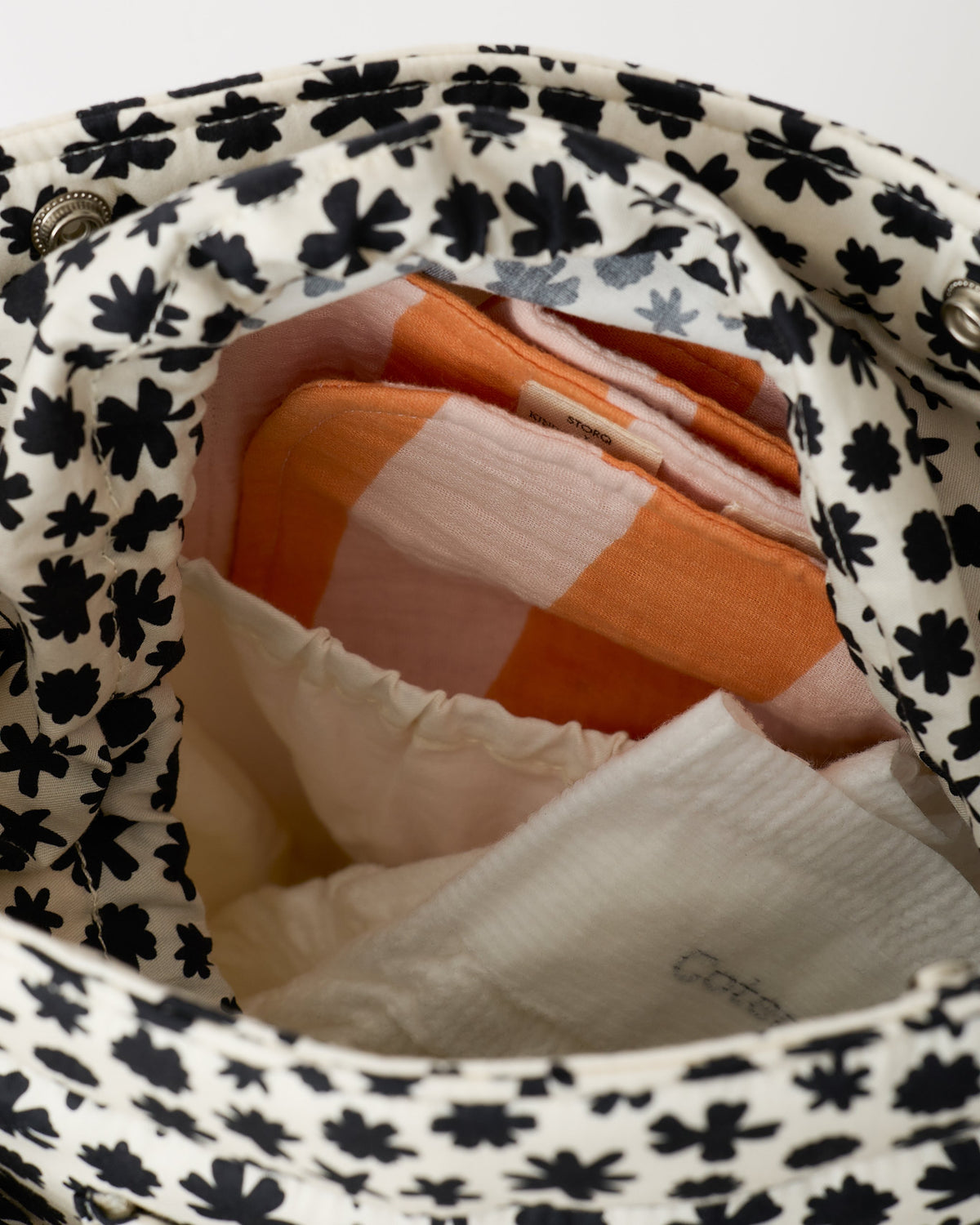 Close up of interior of the Stroller Party™ Bucket Bag in Cream Chamomiles, showing diaper, pocket and Organic Muslin Burp Cloth in Pink Wide Stripe, on a beige background.