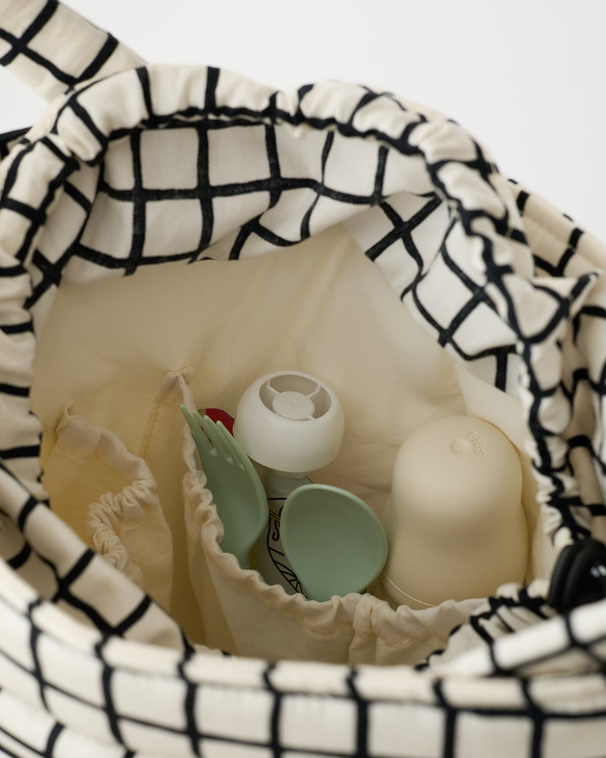 Close up view of interior of Stroller Party™ Bucket Bag in Black Grid including pockets, pouch, milk bottle, utensils and more, on an off white background.