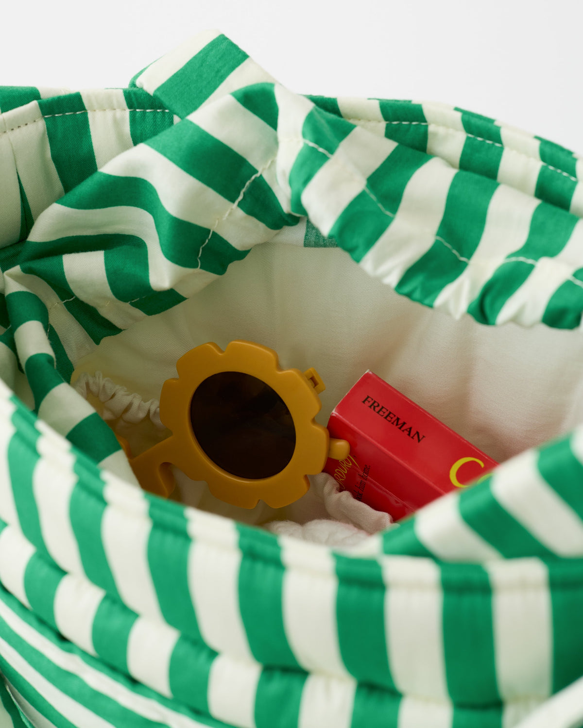 Close up of Stroller Party™ Bucket Bag interior, including pockets, a book, shades and more, on a beige background.