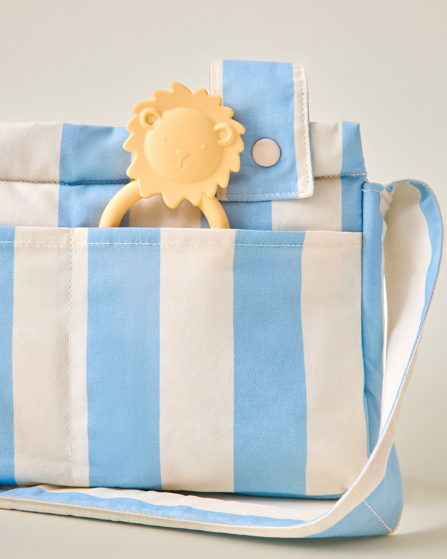 Close-up of Stroller Party Caddy Bag in Blue Wide Stripe pattern with a little lion chew toy sticking out of pocket on a beige background