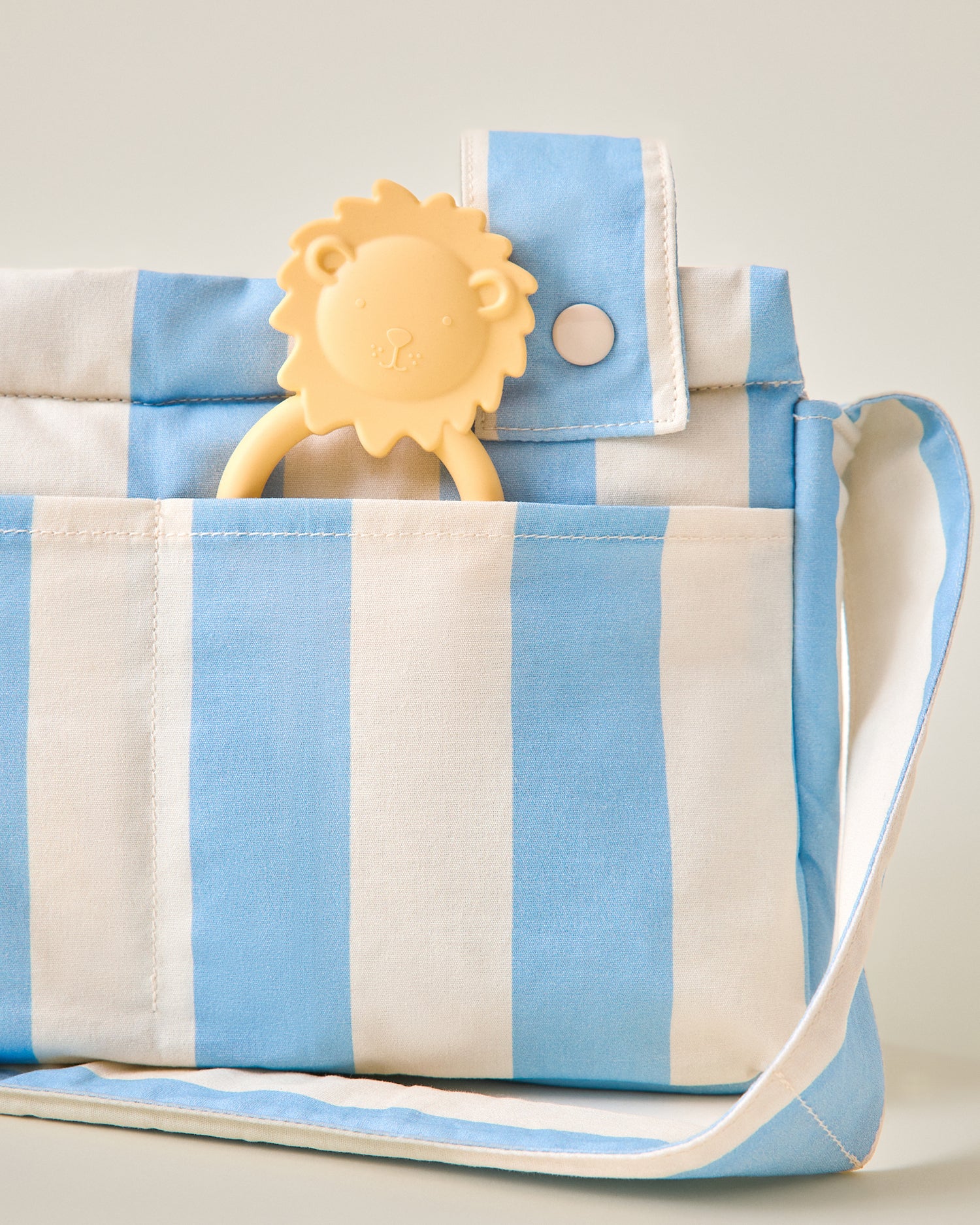 Close-up of Stroller Party Caddy Bag in Blue Wide Stripe pattern with a little lion chew toy sticking out of pocket on a beige background