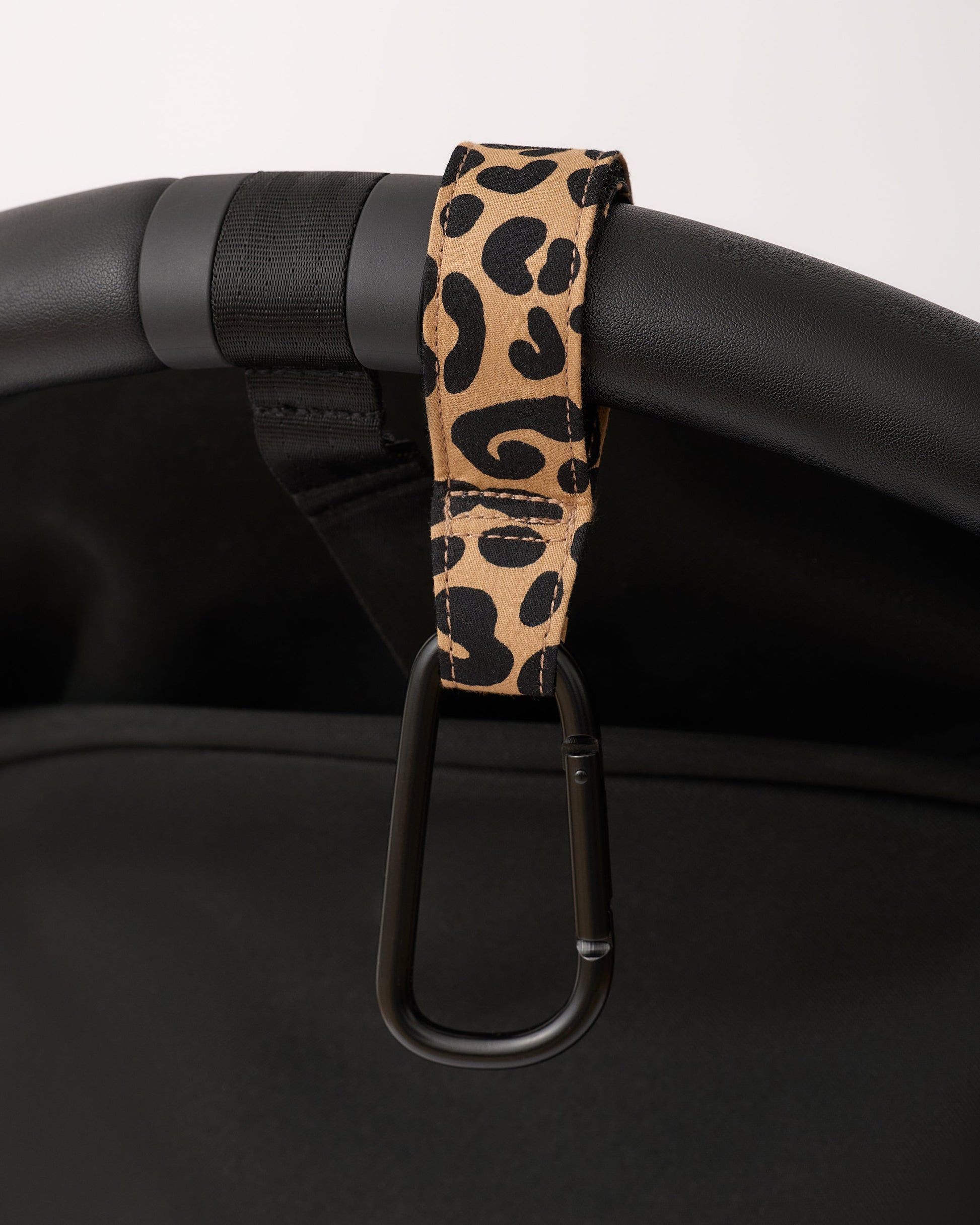 Stroller Party™ Clips (2-Pack) in Leopard hanging from handle bar of black stroller, on a beige background.