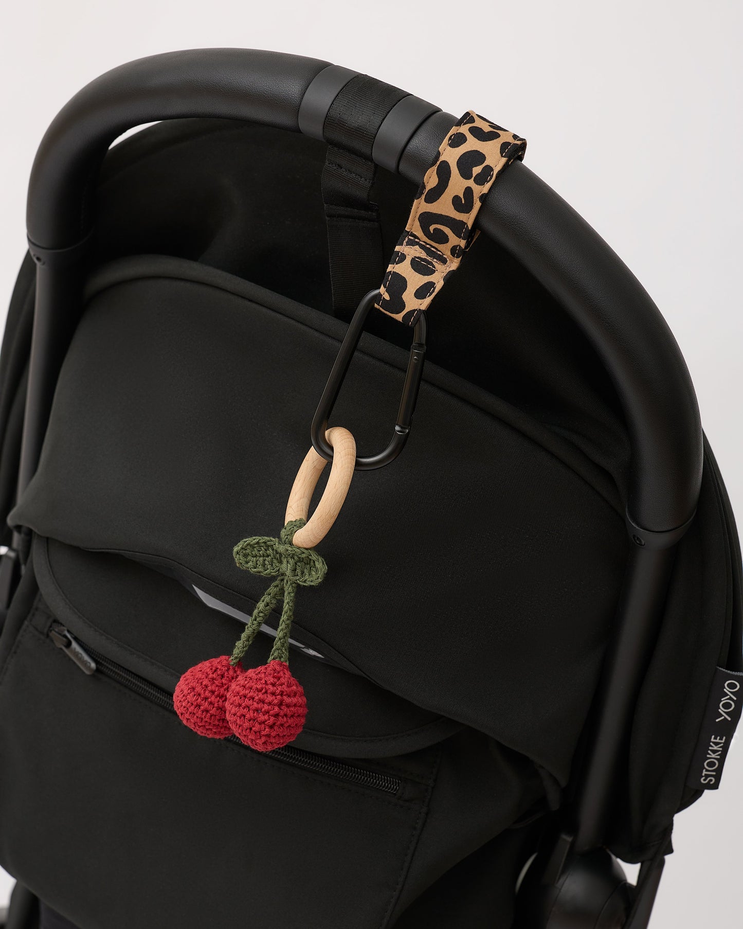 Stroller Party™ Clips (2-Pack) in Leopard hanging from handle bar of black stroller, holding a cute woven cherry chew toy, on a beige background.