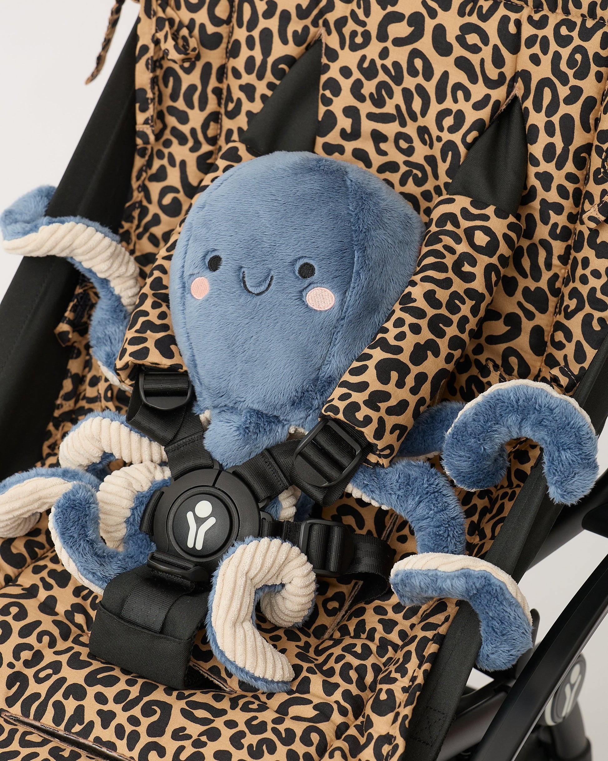 Close up of an octopus lovey all buckled into black stroller with Stroller Party™ Seat Liner & Strap Covers in Leopard, on a beige background.