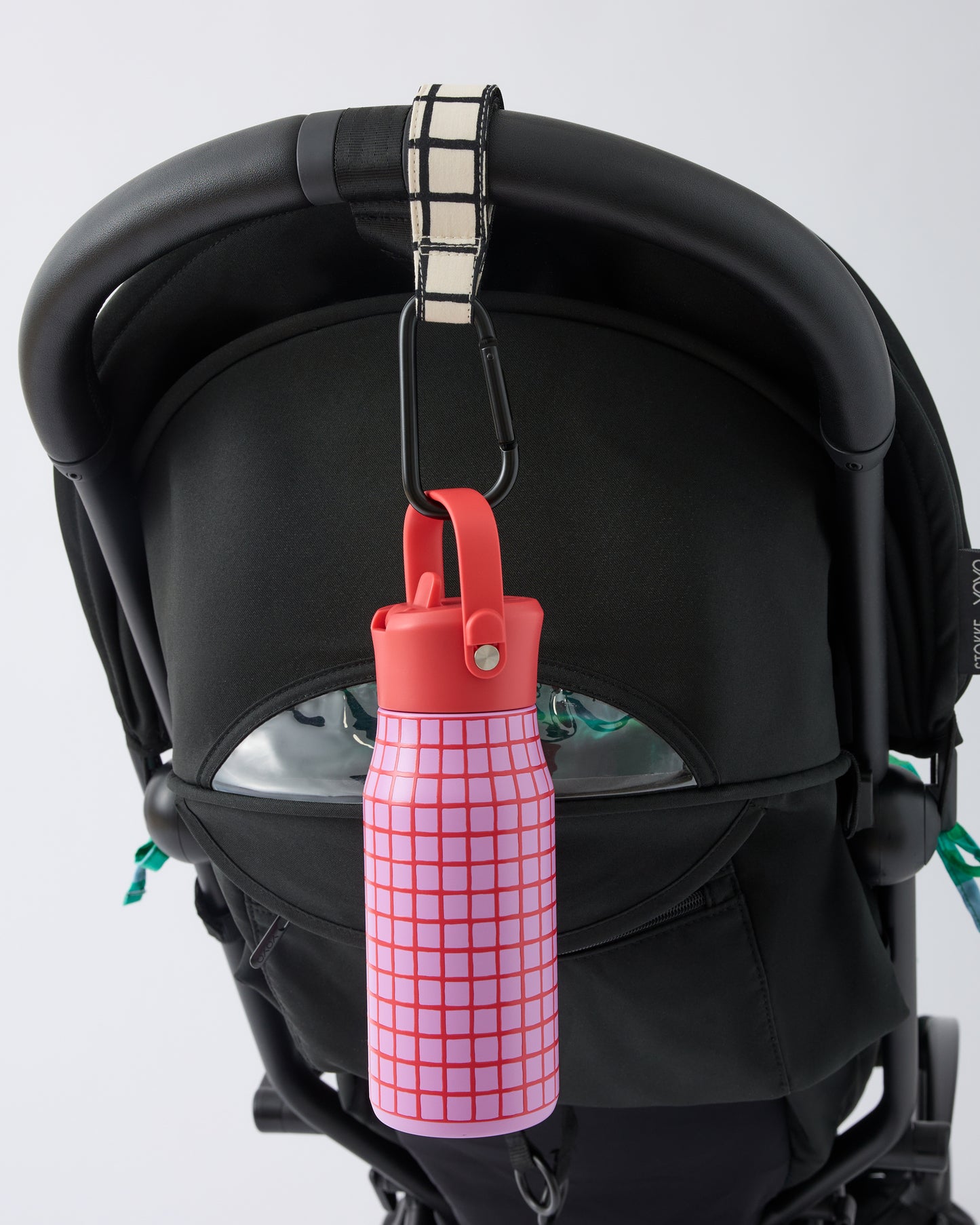 Black stroller handle bar with Stroller Party™ Clips (2-Pack) in Black Grid attached, while holding a Cool Kid Water Bottle in Orchid Grid, on a beige background.