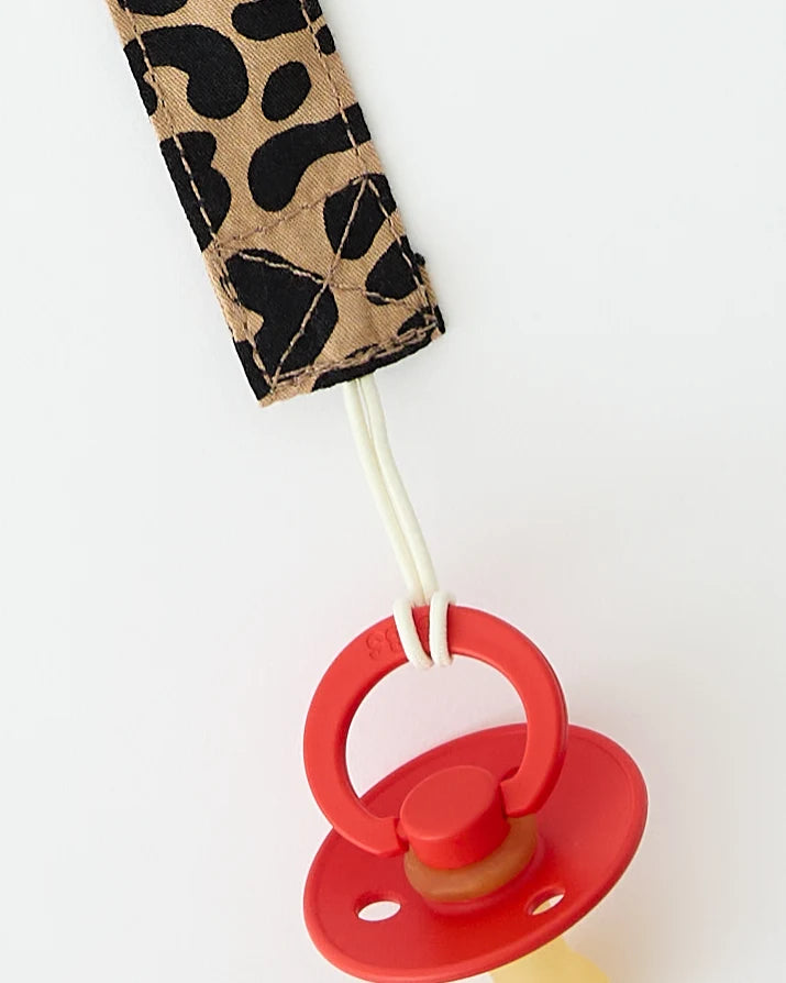 Close up of Pacifier Clip in Leopard, holding onto a red pacifier, on a beige background.