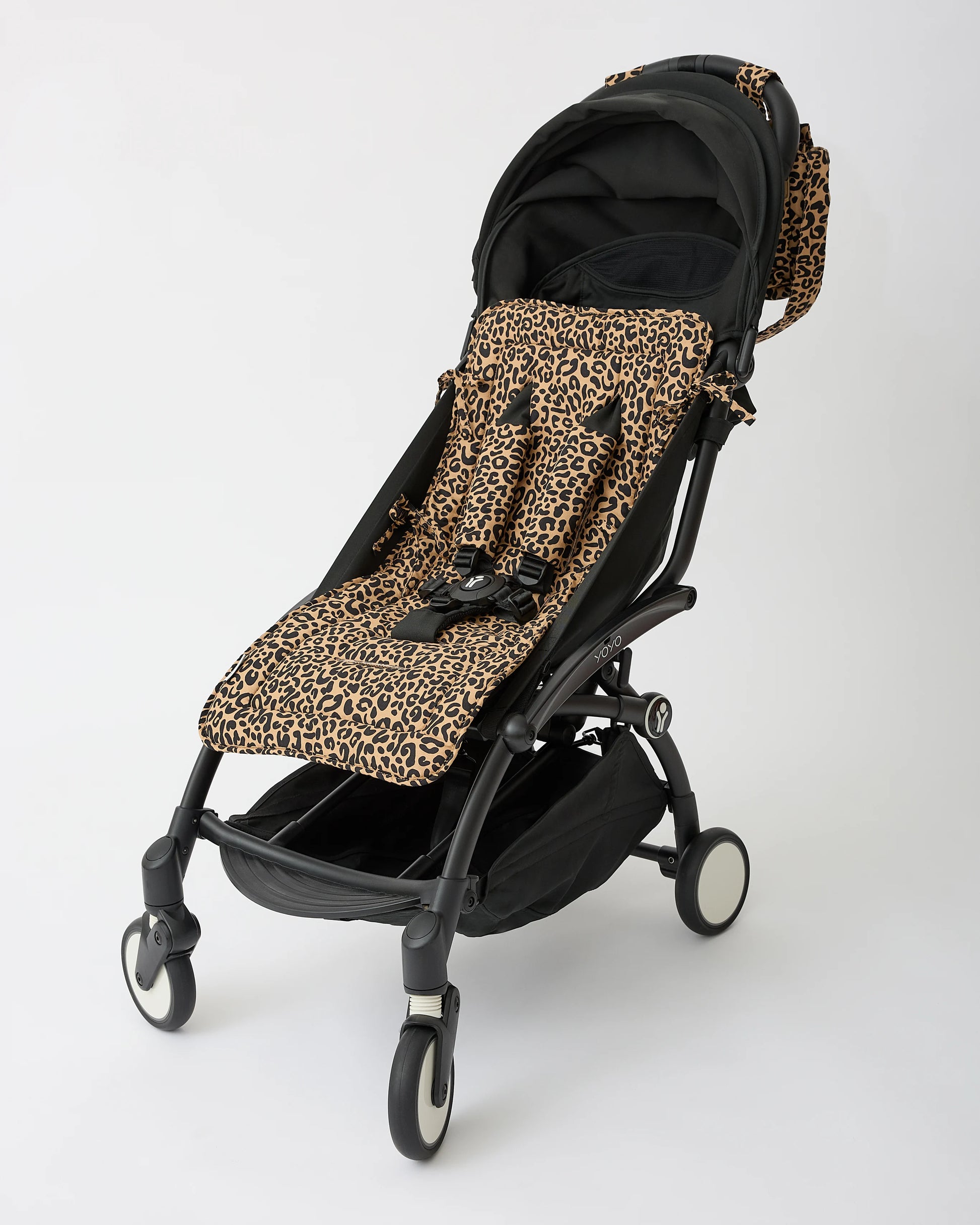 A black stroller with Stroller Party™ Seat Liner & Strap Covers in Leopard, secured with a black safety harness on a black stroller frame, and Stroller Party™ Caddy Bag in Leopard attached to handle bar, on a beige background.