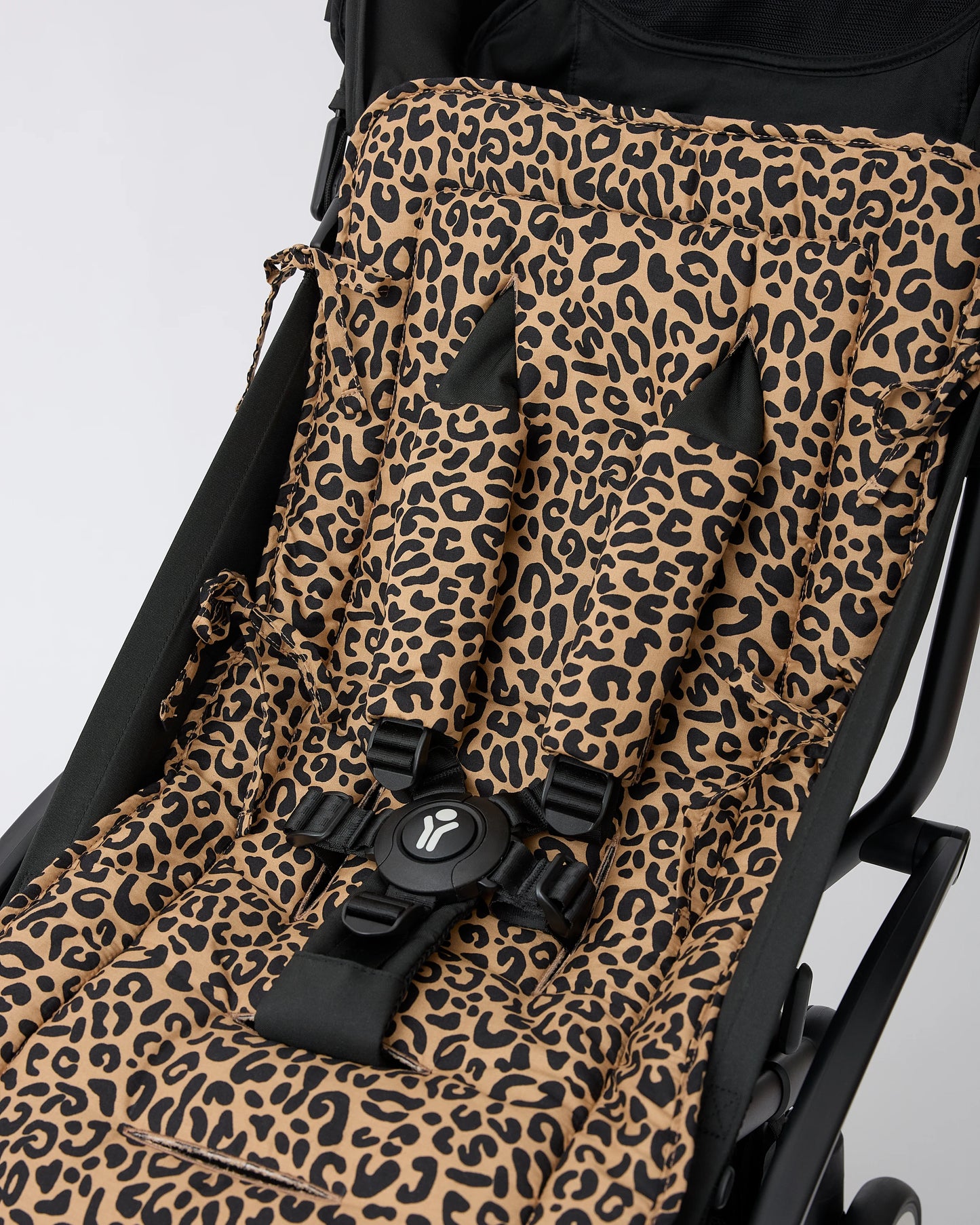 A close-up of a black stroller with Stroller Party™ Seat Liner & Strap Covers in Leopard, secured with a black safety harness on a black stroller frame, on a beige background.