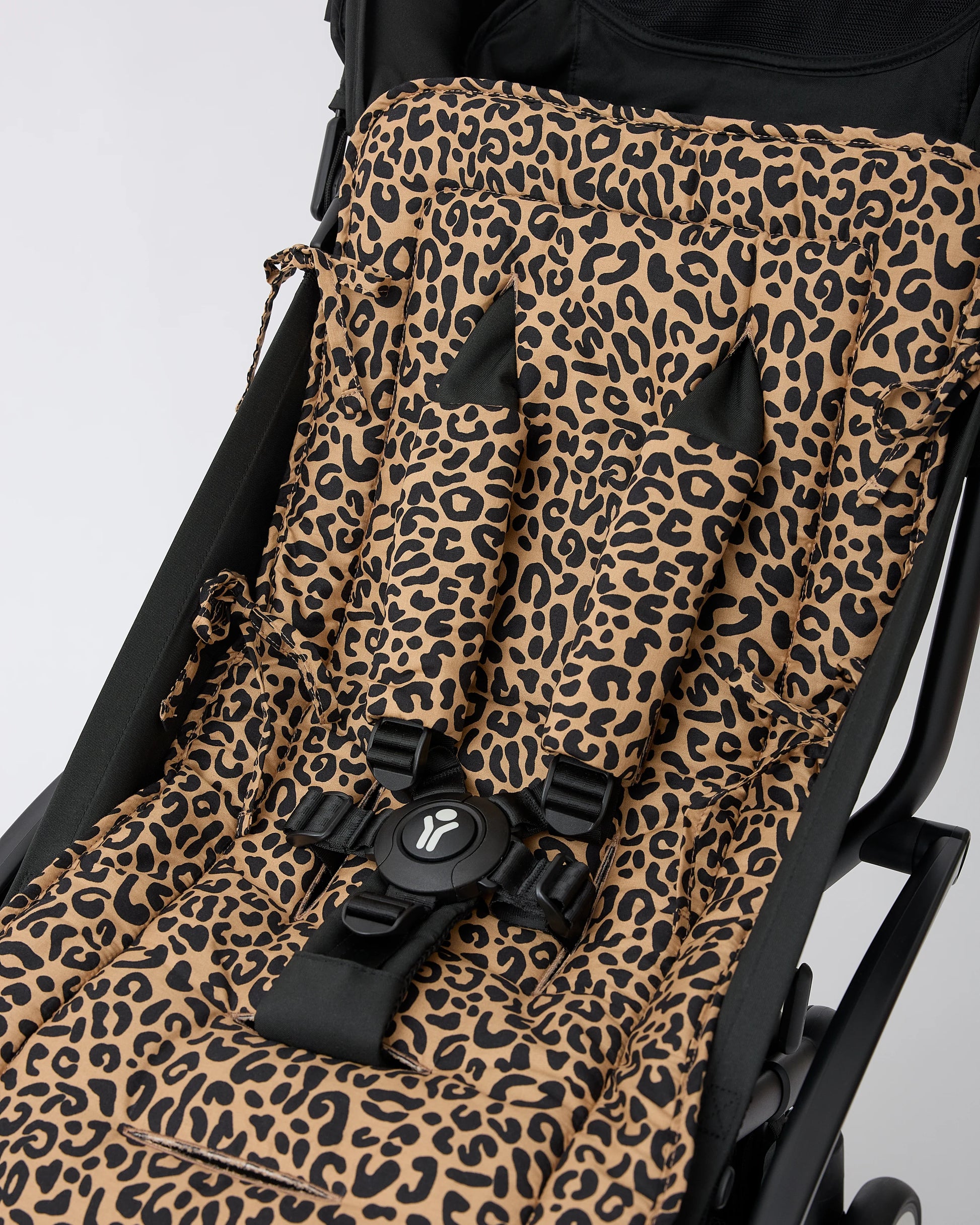 A close-up of a black stroller with Stroller Party™ Seat Liner & Strap Covers in Leopard, secured with a black safety harness on a black stroller frame, on a beige background.