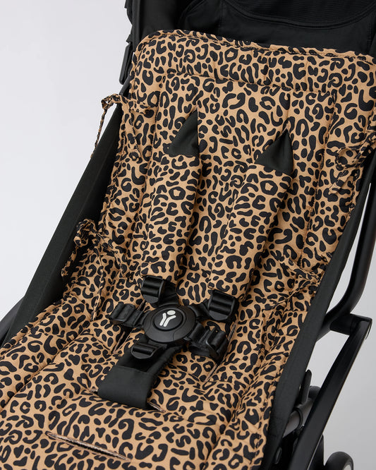 A close-up of a black stroller with Stroller Party™ Seat Liner & Strap Covers in Leopard, secured with a black safety harness on a black stroller frame, on a beige background.