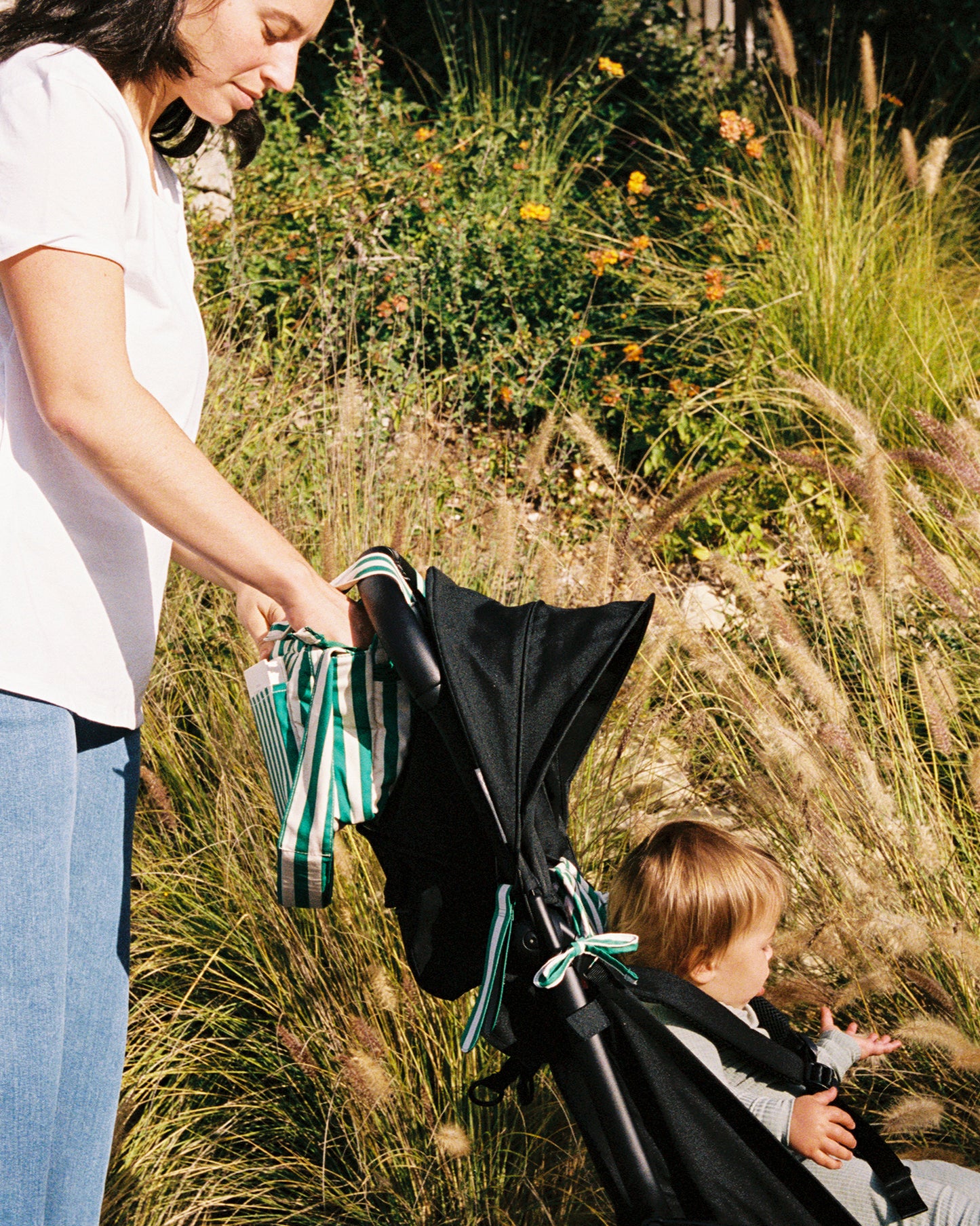 Woman pushing a stroller with Stroller Party accessories in Green Stripe, with a child inside, surrounded by tall grass and wildflowers.