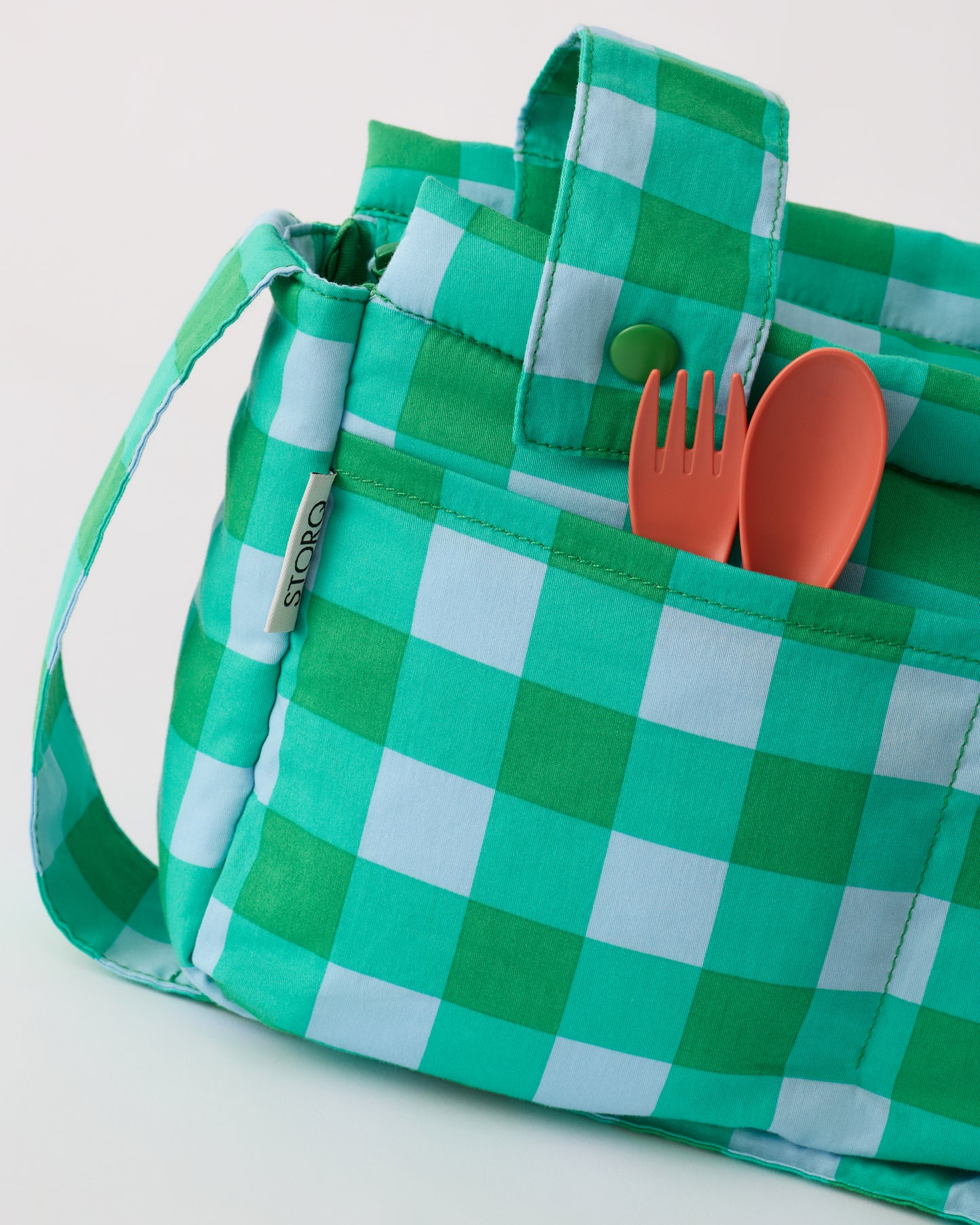 Close up of pockets of the Stroller Party™ Caddy Bag in Teal Gingham, holding orange spoon and fork, on a beige background.