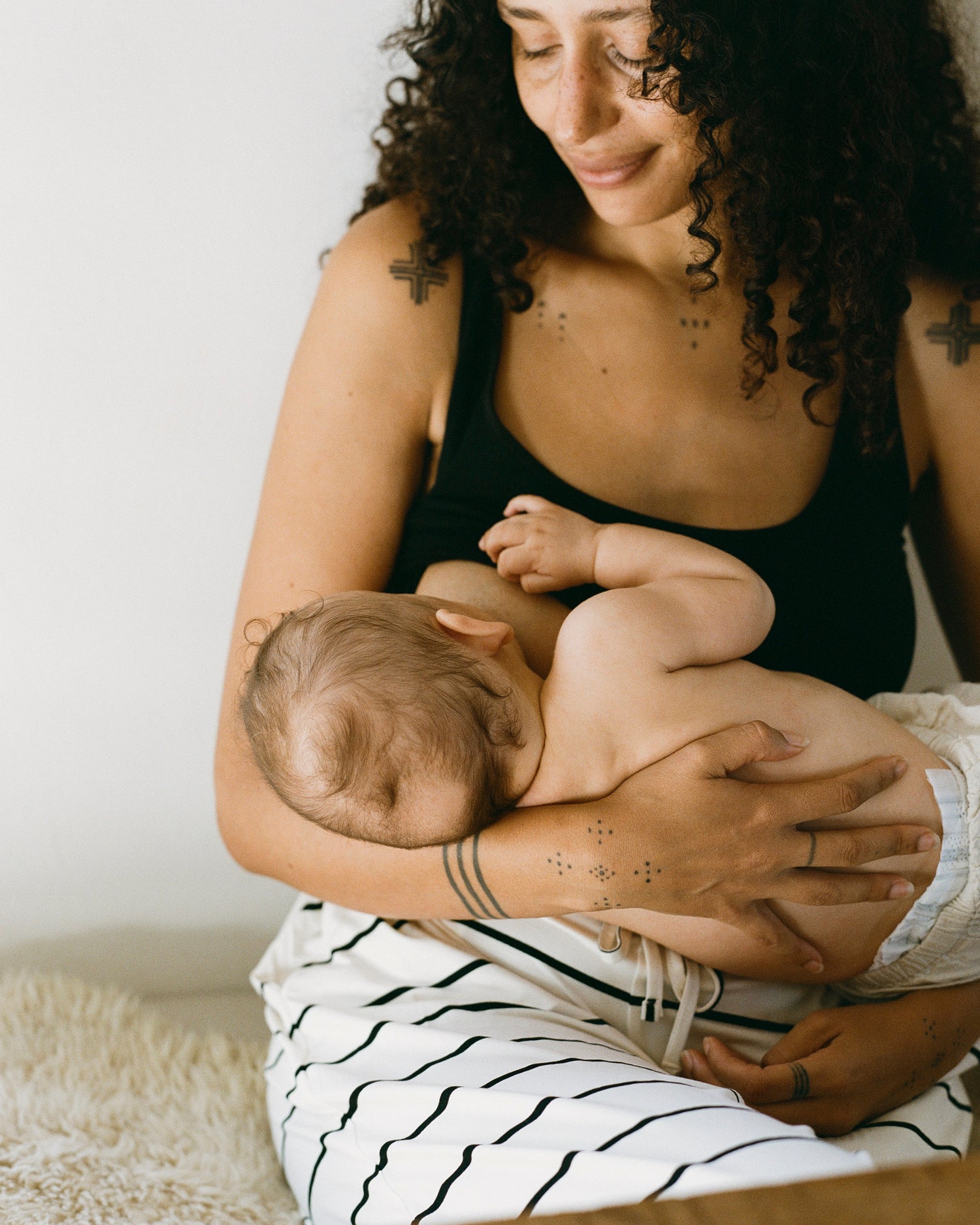 Model wearing Sublime® Bamboo Scoop Neck Maternity & Nursing Longline Bra Top in Black, nursing a baby@model_info:Natasha is wearing a Medium.