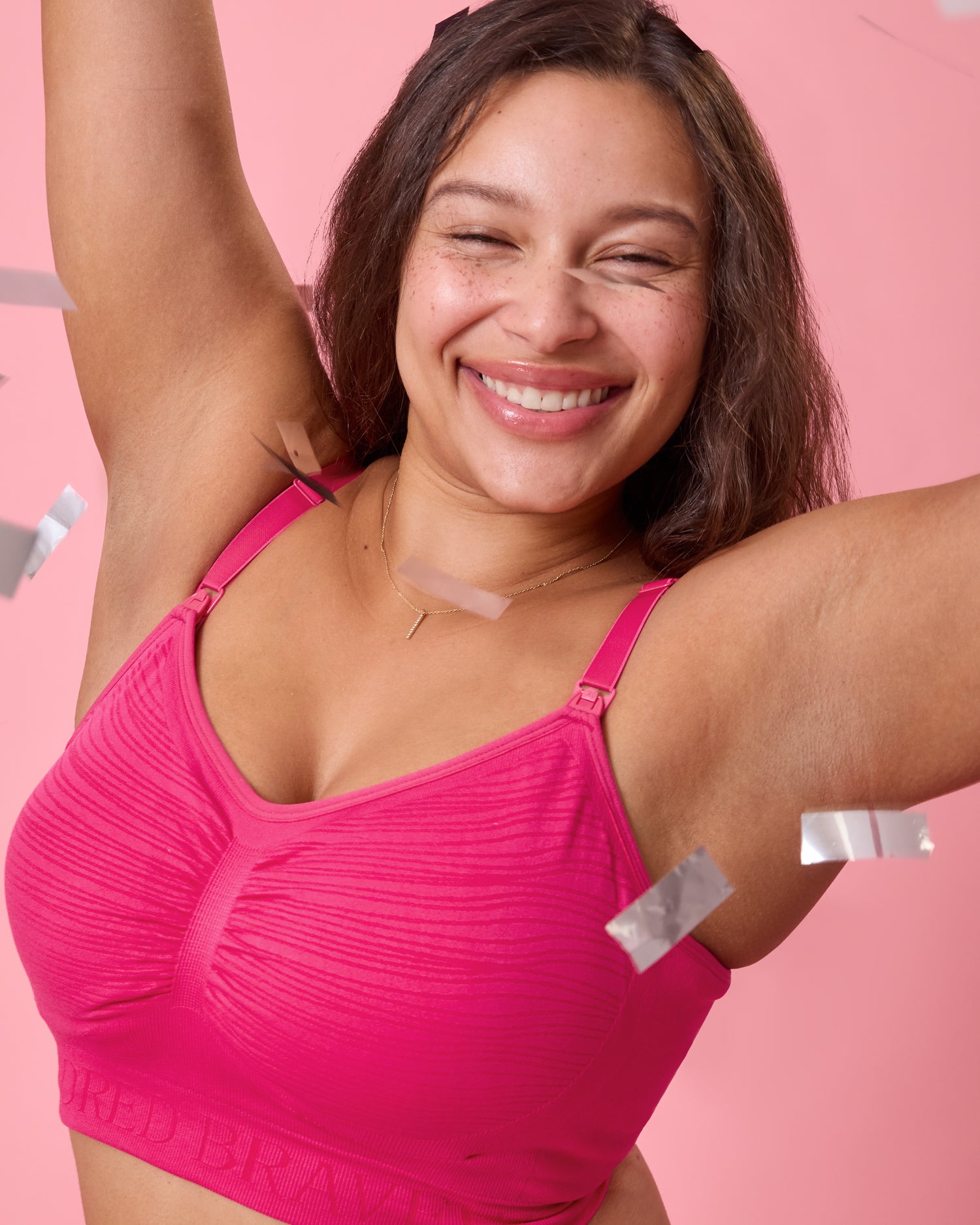 Front view of model wearing Sublime Hands-Free Pumping & Nursing Bra  Raspberry Pink on a pink background smiling happily and with confetti falling around