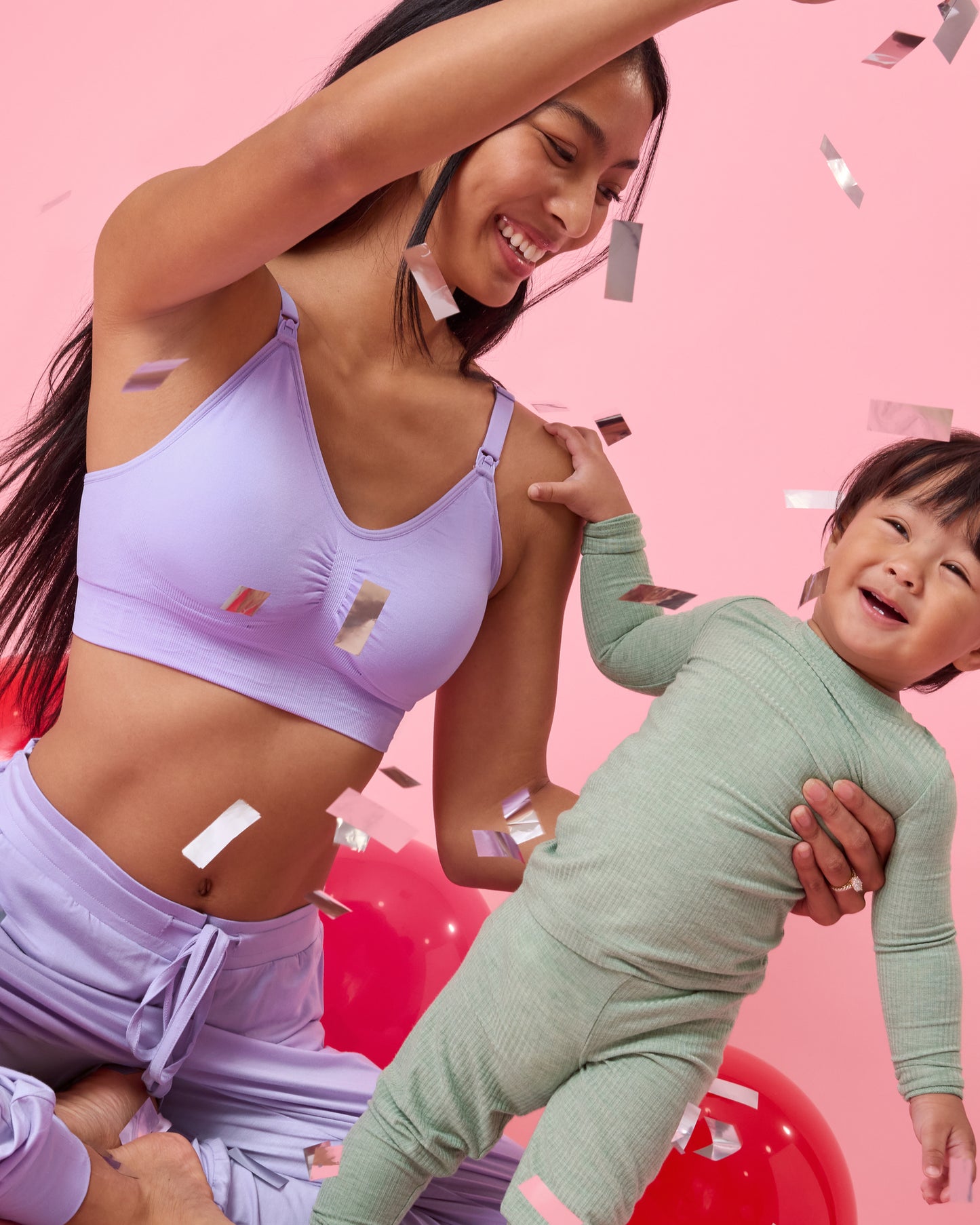 Front view of model wearing Simply Sublime Nursing Bra in Lavender on a pink background sitting on the floor with a baby and laughing happily while gently dropping confetti around them