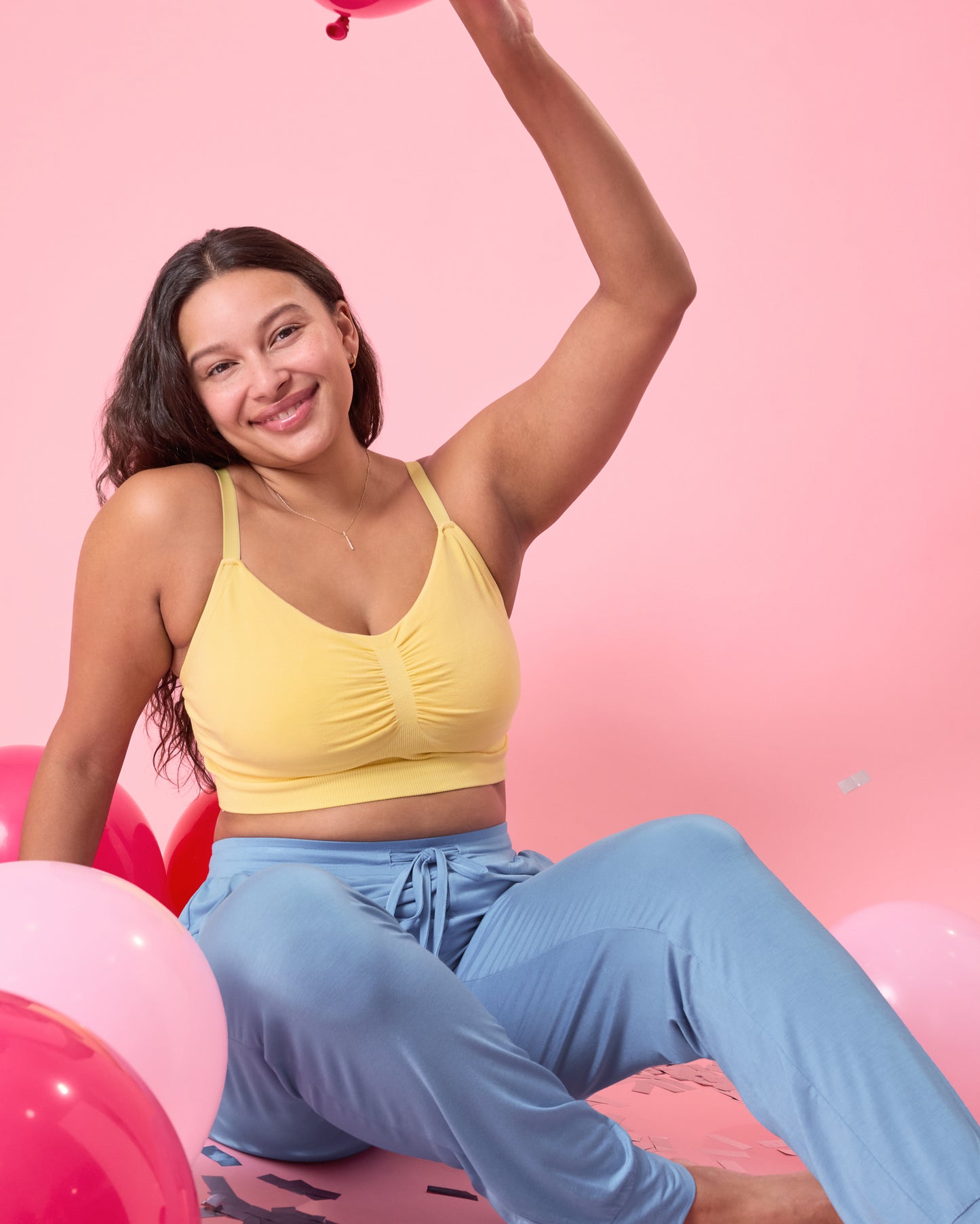 Model wearing Sublime Bamboo Hands-Free Pumping Lounge & Sleep Bra in Dandelion sitting in a relaxed pose on the floor surrounded by pink balloons and in front of a pink background